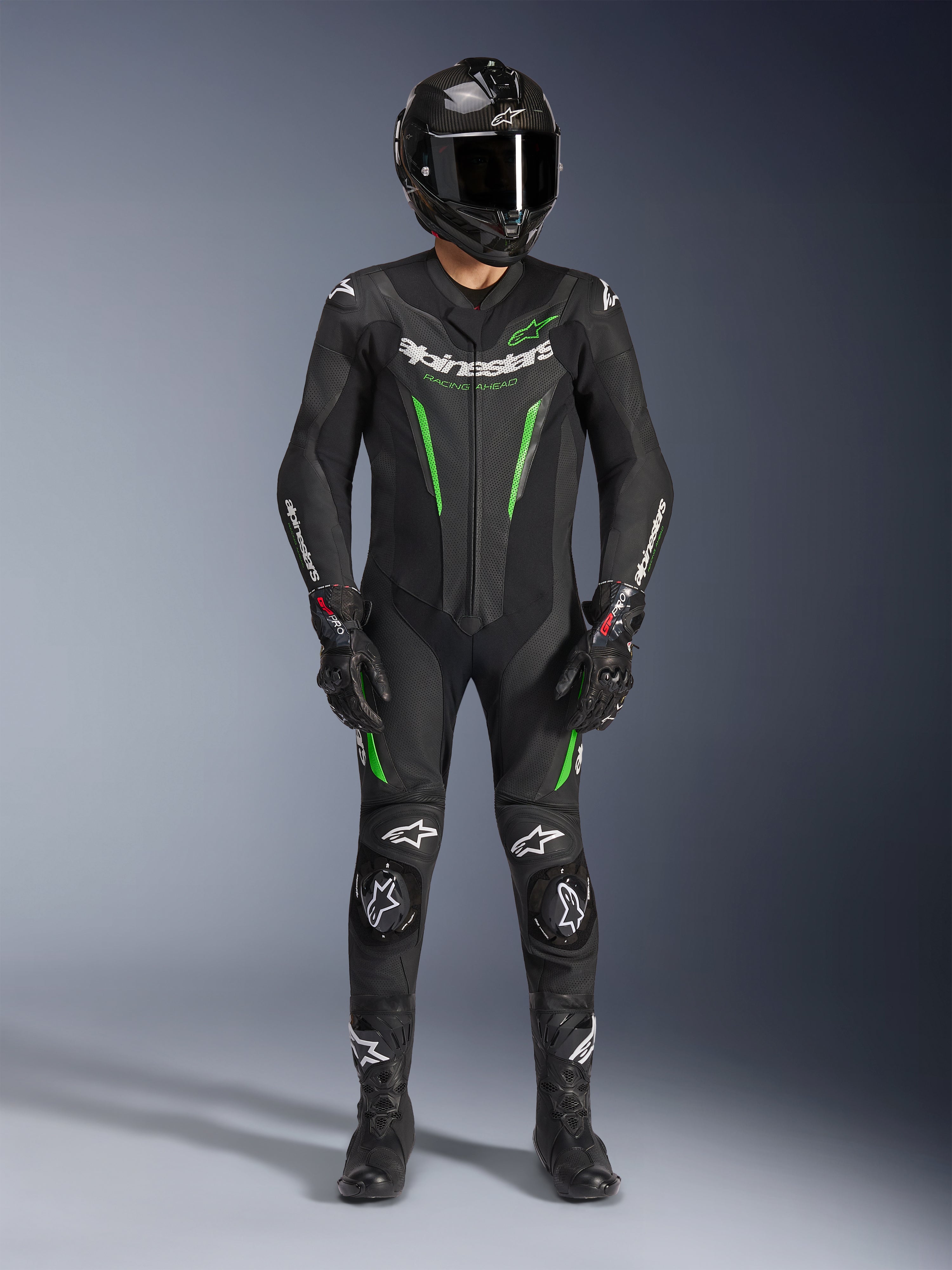 Motorcycle racer wearing Alpinestars GP Force V2 Leather Suit - 1Piece in Black and Green, paired with a carbon fiber helmet and racing gloves, featuring perforated leather panels and white Alpinestars logos, professional track and sport riding gear