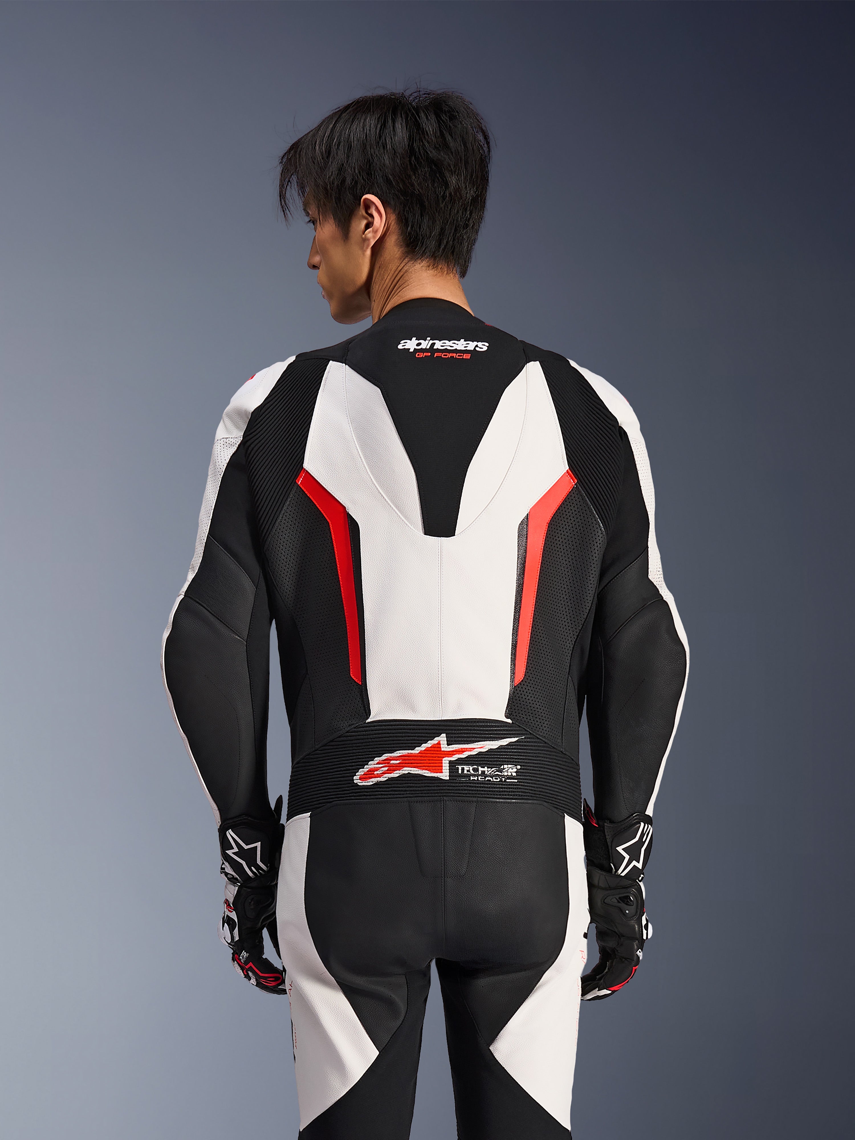 Alpinestars GP Force V2 Leather Suit 1Piece, White Black and Red Fluo, back view on male model, featuring aerodynamic speed hump, perforated leather panels, and Tech-Air Ready logo on the lower back, professional racing gear with black and white gloves