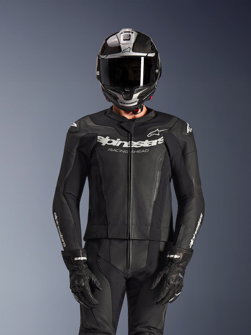 Rider wearing Alpinestars GP Force V2 2-Piece Leather Suit in Black, perforated leather construction with white 'Racing Ahead' chest logo, silver and carbon fiber full-face helmet, black gloves, professional sport and track riding gear