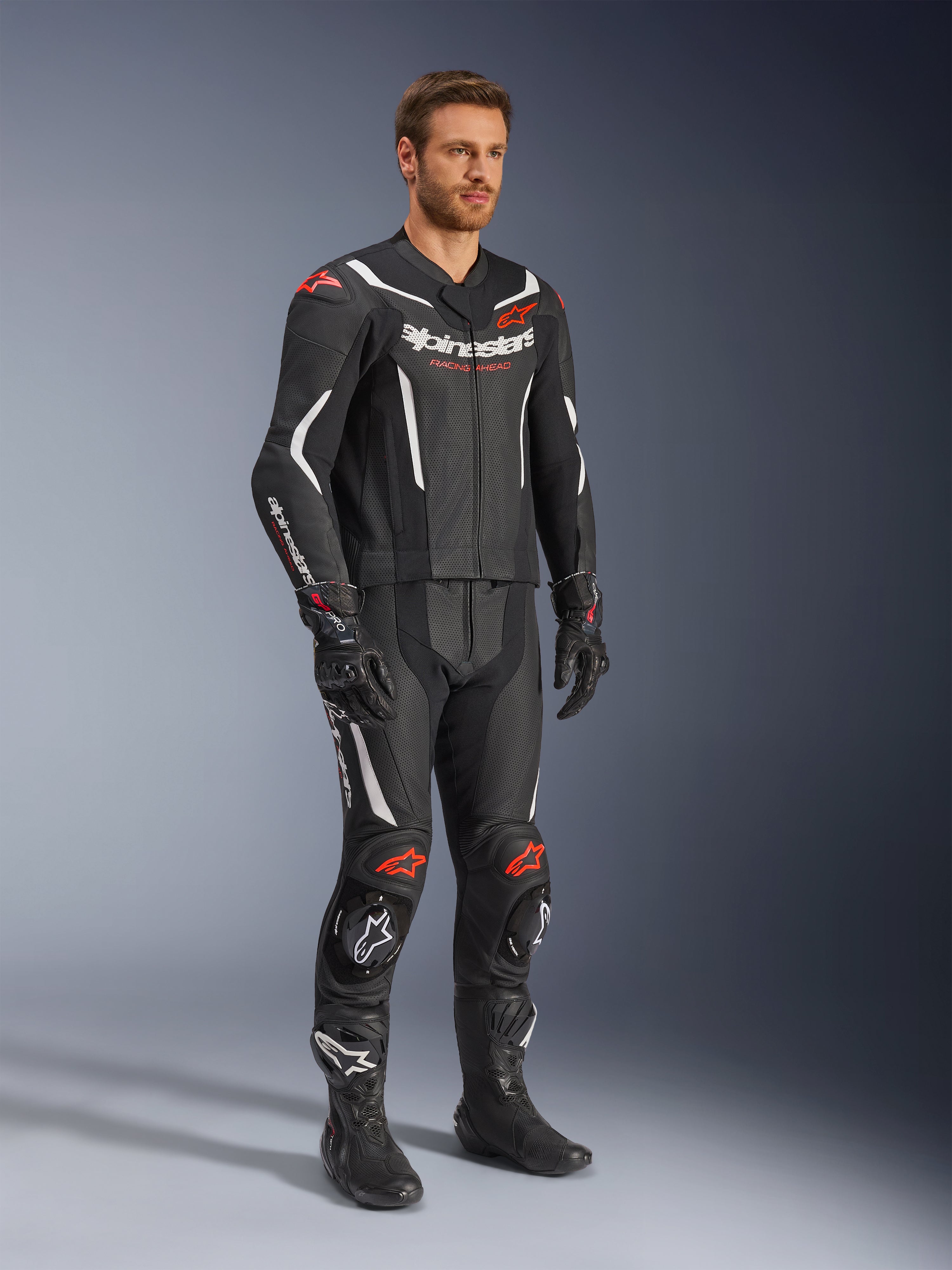 Alpinestars GP Force V2 2-Piece Leather Suit, Black White and Red Fluo, worn by a model with black racing boots and gloves, featuring perforated leather panels, white aerodynamic accents, and red fluo logos on shoulders and knees, professional motorcycle racing gear