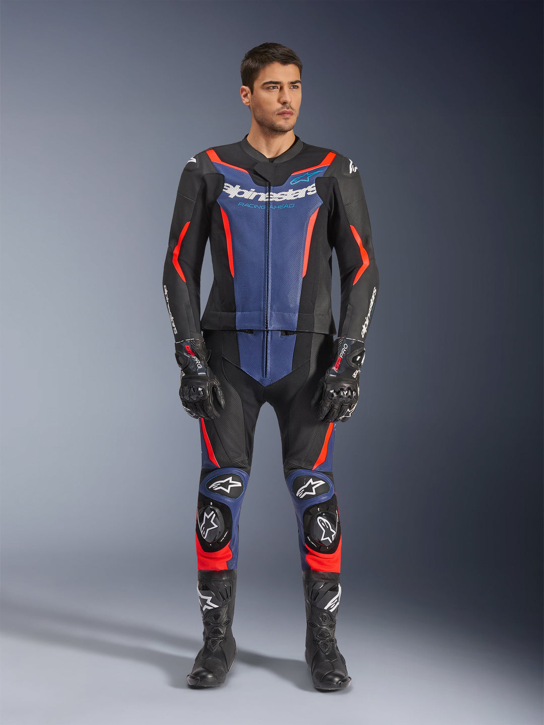 Alpinestars GP Force V2 Leather Suit 2-Piece in Black Blue and Red Fluo, featuring blue panels on the chest and lower legs with vibrant red fluorescent highlights, worn by a male model with black racing boots and gloves, professional motorsport protective apparel