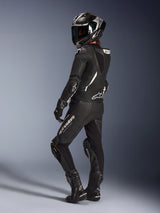Female rider wearing the Alpinestars Stella GP-R7 2-Piece Leather Suit in Black, full-face racing helmet with carbon finish, matching gloves and boots, back view showcasing the aerodynamic hump and white Alpinestars logos, professional track and sport riding gear