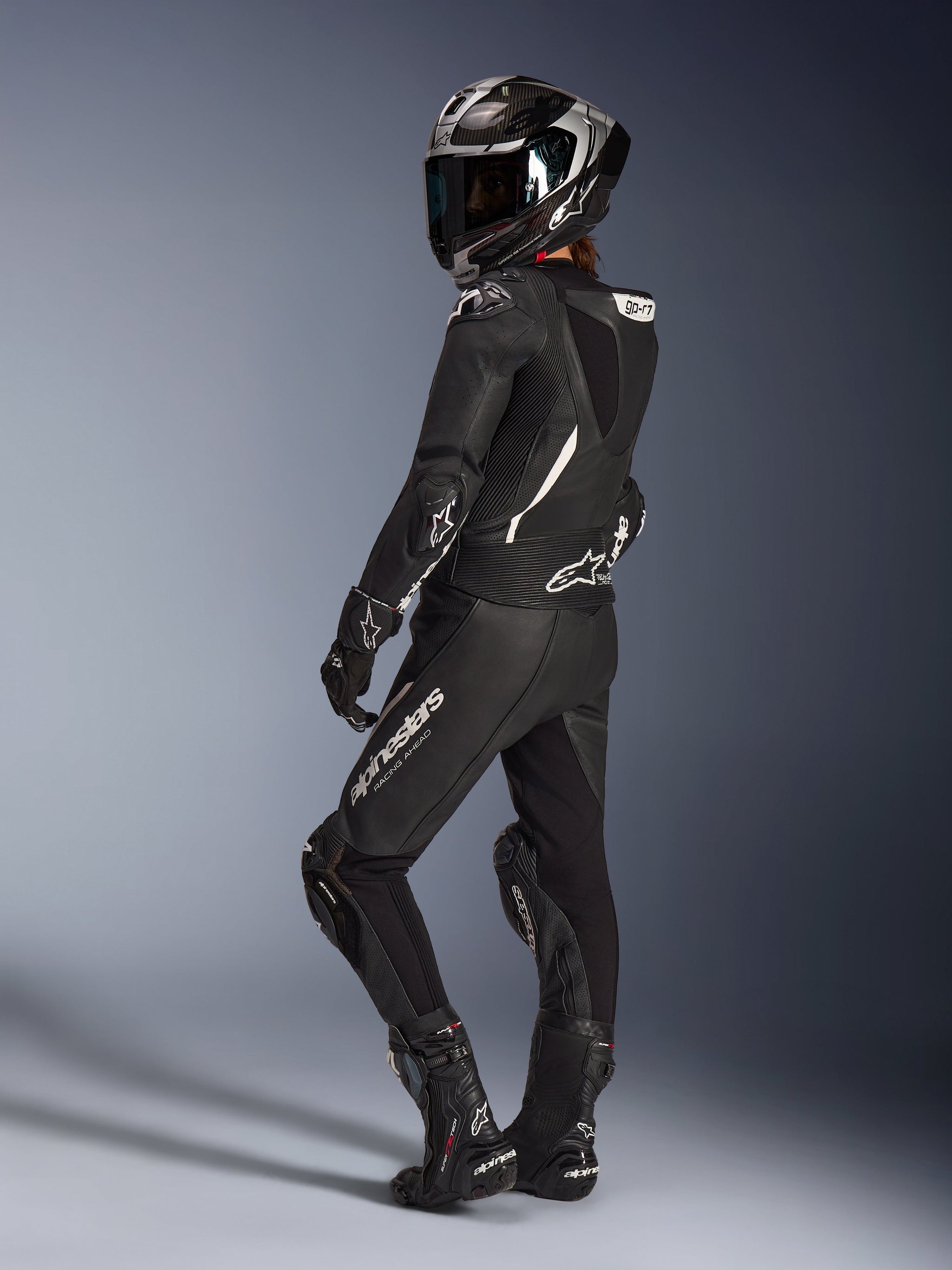 Female rider wearing the Alpinestars Stella GP-R7 2-Piece Leather Suit in Black, full-face racing helmet with carbon finish, matching gloves and boots, back view showcasing the aerodynamic hump and white Alpinestars logos, professional track and sport riding gear
