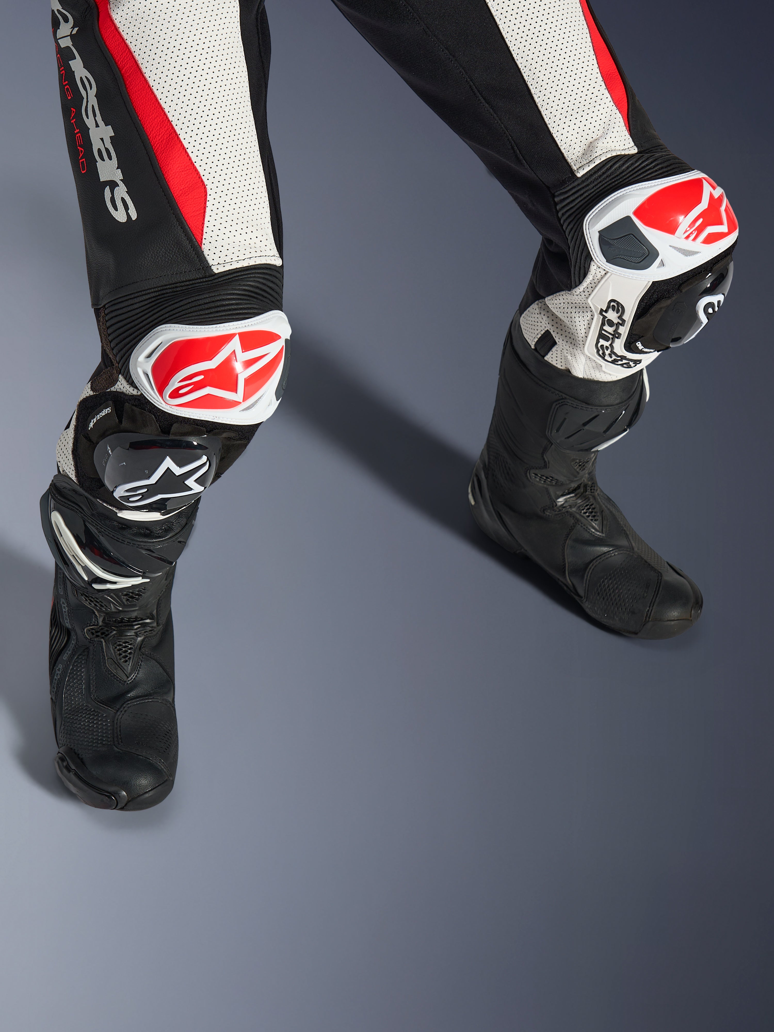 Alpinestars Woman Stella GP-R7 2-Piece Leather Suit, Black White and Red Fluo, close-up of legs and knees featuring red and white GP DFS RR sliders, perforated leather construction with accordion stretch panels, worn with black performance racing boots