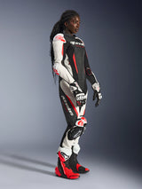 Female rider wearing the Alpinestars Stella GP-R7 2-Piece Leather Suit in Black, White, and Red Fluo, paired with matching racing gloves and boots, featuring perforated leather panels and protective armor on shoulders, elbows, and knees, standing in a studio setting.