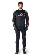 Male model wearing the Alpinestars T-SPS V2 Waterproof Jacket in Black White Red Fluo, paired with black motorcycle pants and gloves, featuring a white Alpinestars logo on the chest and red accents on the sleeves.
