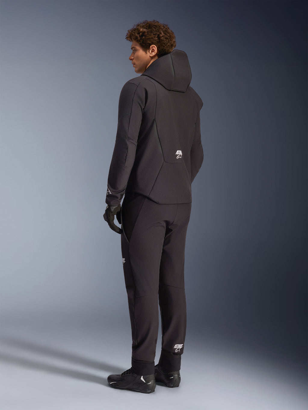 Alpinestars Aeroshell Waterproof Jacket, Black softshell motorcycle hoodie, back view on model, worn with matching black pants and riding shoes, featuring a hood and small white Alpinestars logo on the back, sleek waterproof gear for road and casual touring