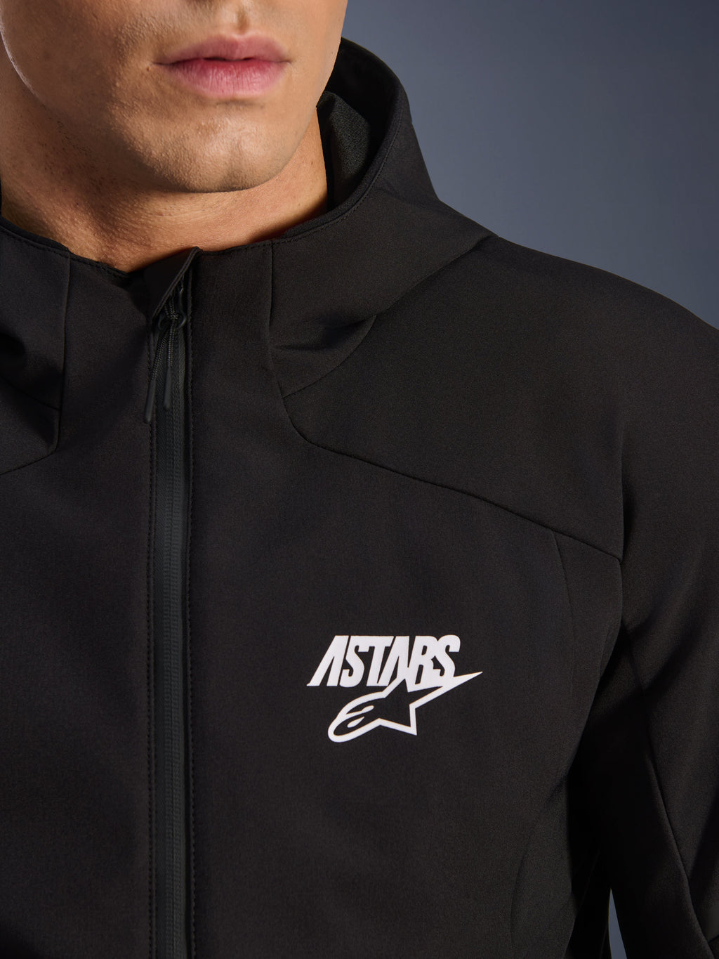 Alpinestars Aeroshell Waterproof Jacket, black motorcycle softshell jacket, close-up view of the chest featuring a white ASTARS star logo and waterproof front zipper, integrated hood, modern sport-styled design
