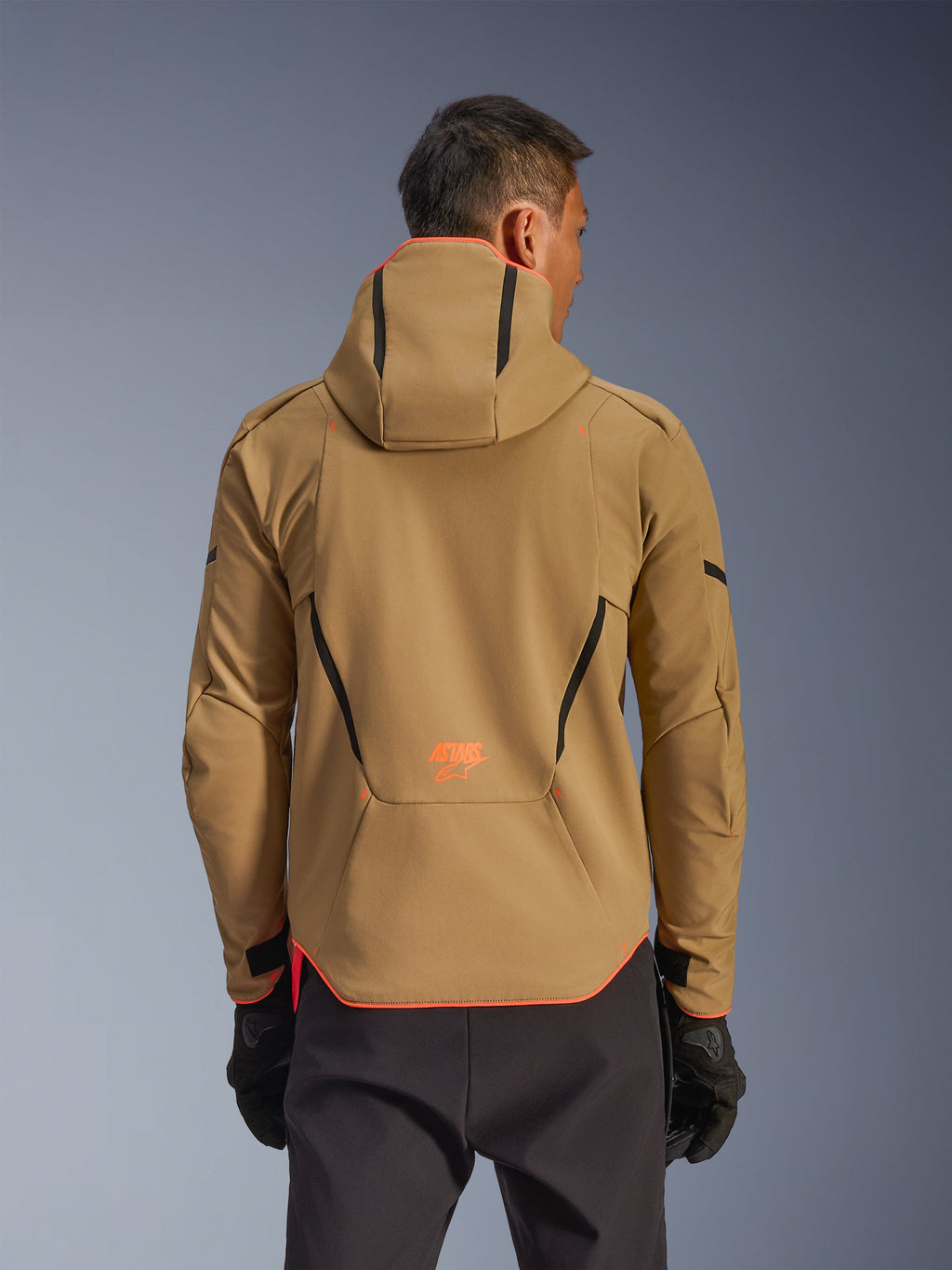 Alpinestars Aeroshell Waterproof Jacket, Dark Khaki and Orange Fluo, rear view showing hoodie design with orange trim on the hood and hem, orange logo on lower back, worn with black gloves, crossover sports softshell motorcycle gear.