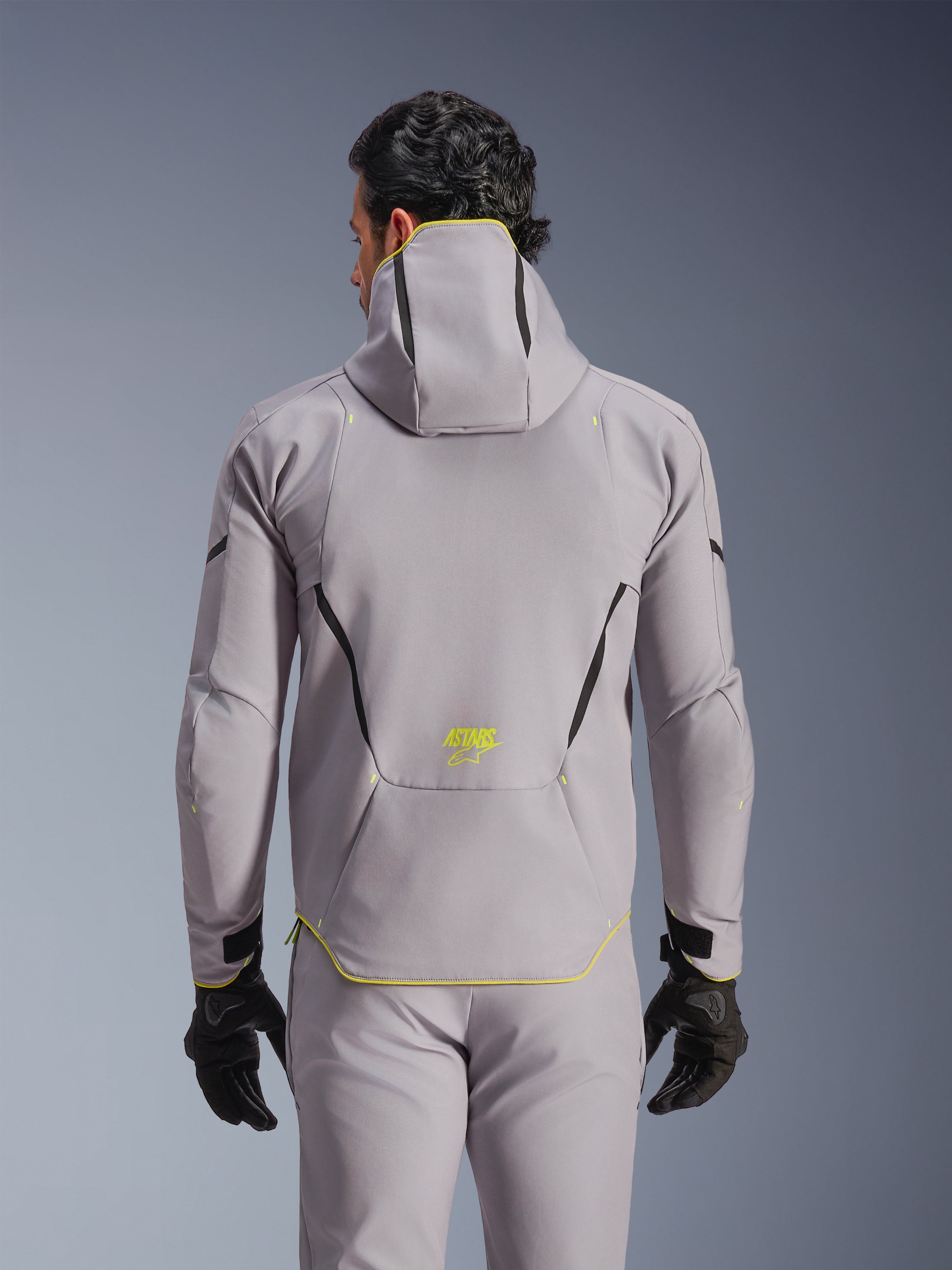 Alpinestars Aeroshell Waterproof Jacket, Mid Gray with Yellow Fluo accents, back view showing integrated hood and black contrast lines, yellow ASTARS logo on lower back, worn with matching gray pants and black gloves, sport softshell motorcycle jacket