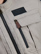 Alpinestars Borrego Drystar® Jacket, Warm Gray, close-up detail of chest area featuring a black Alpinestars branding patch, chest pocket with flap, and a vertical zipper with a brown leather pull tab, durable gray textile canvas construction.