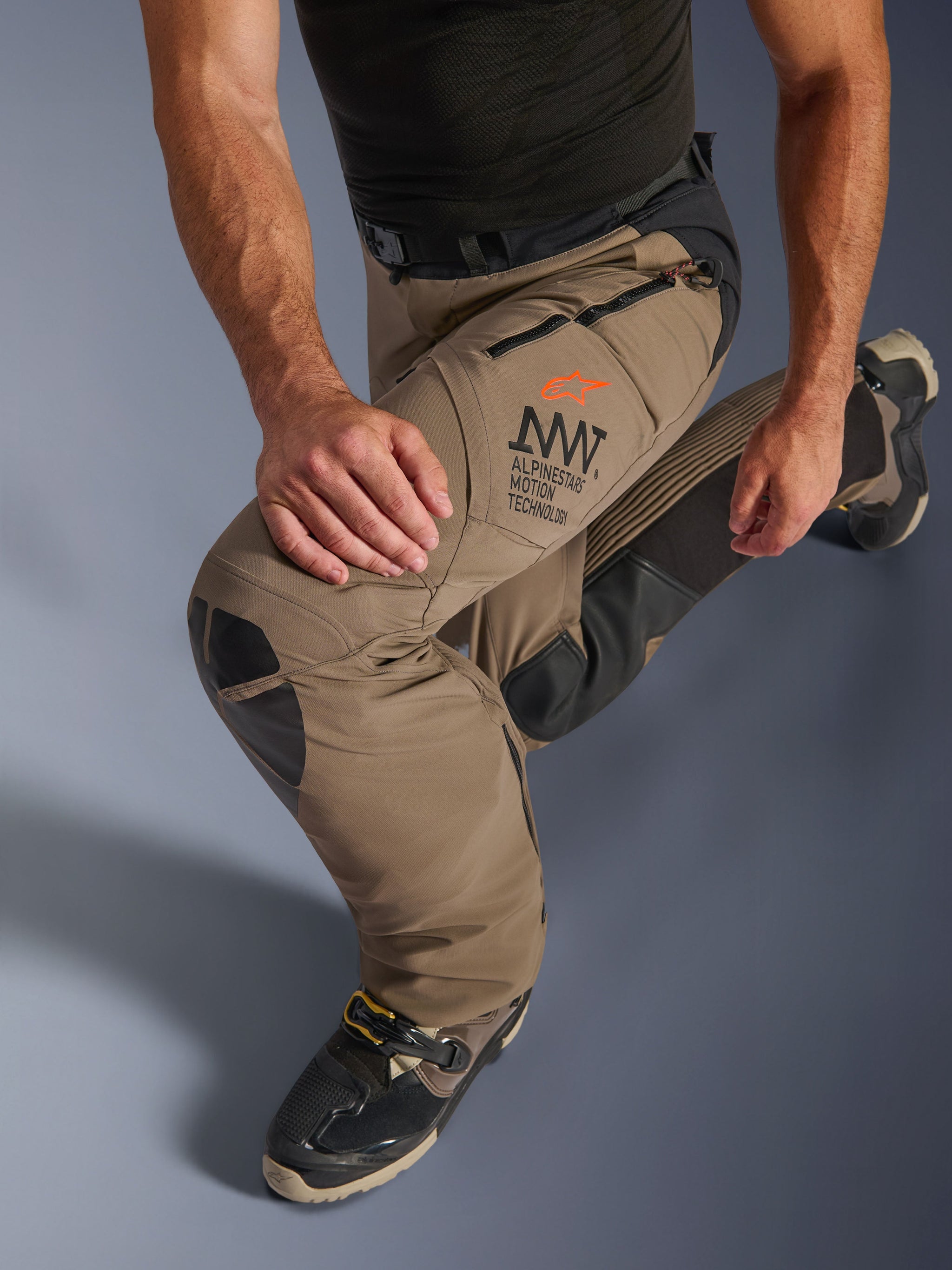 Alpinestars AMT-8 Stretch Drystar® XF Pants in Walnut Black, featuring Alpinestars Motion Technology (AMT) branding on the thigh, long zippered ventilation ports, and durable leather knee panels, shown on a rider in a kneeling position to highlight flexibility.
