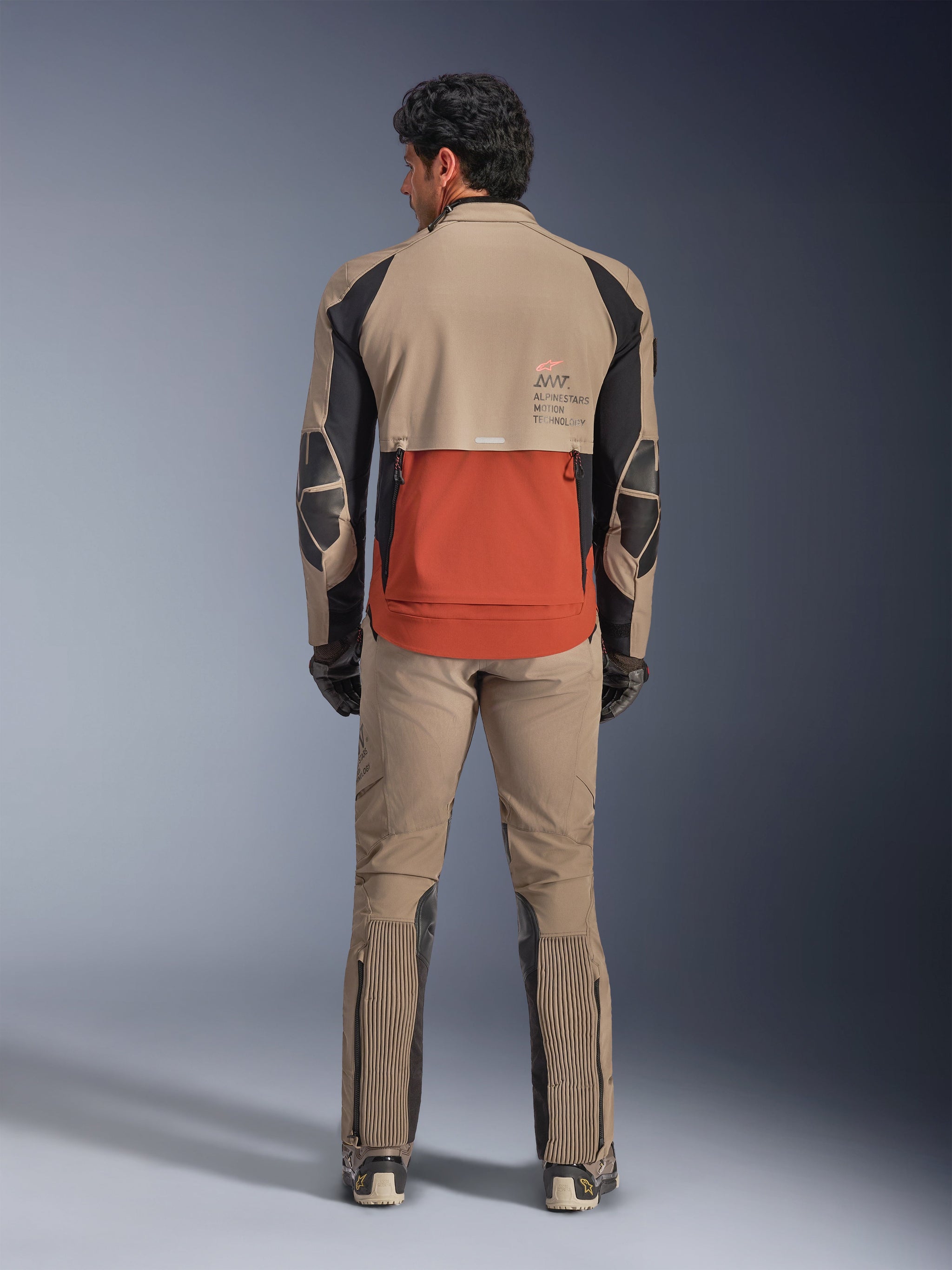 Alpinestars AMT-8 Stretch Drystar® XF Jacket, Walnut Rust Black, rear view on a rider with matching adventure pants and gloves, featuring a prominent rust-colored rear panel, walnut upper sections, and black stretch accents, designed for long-range touring.