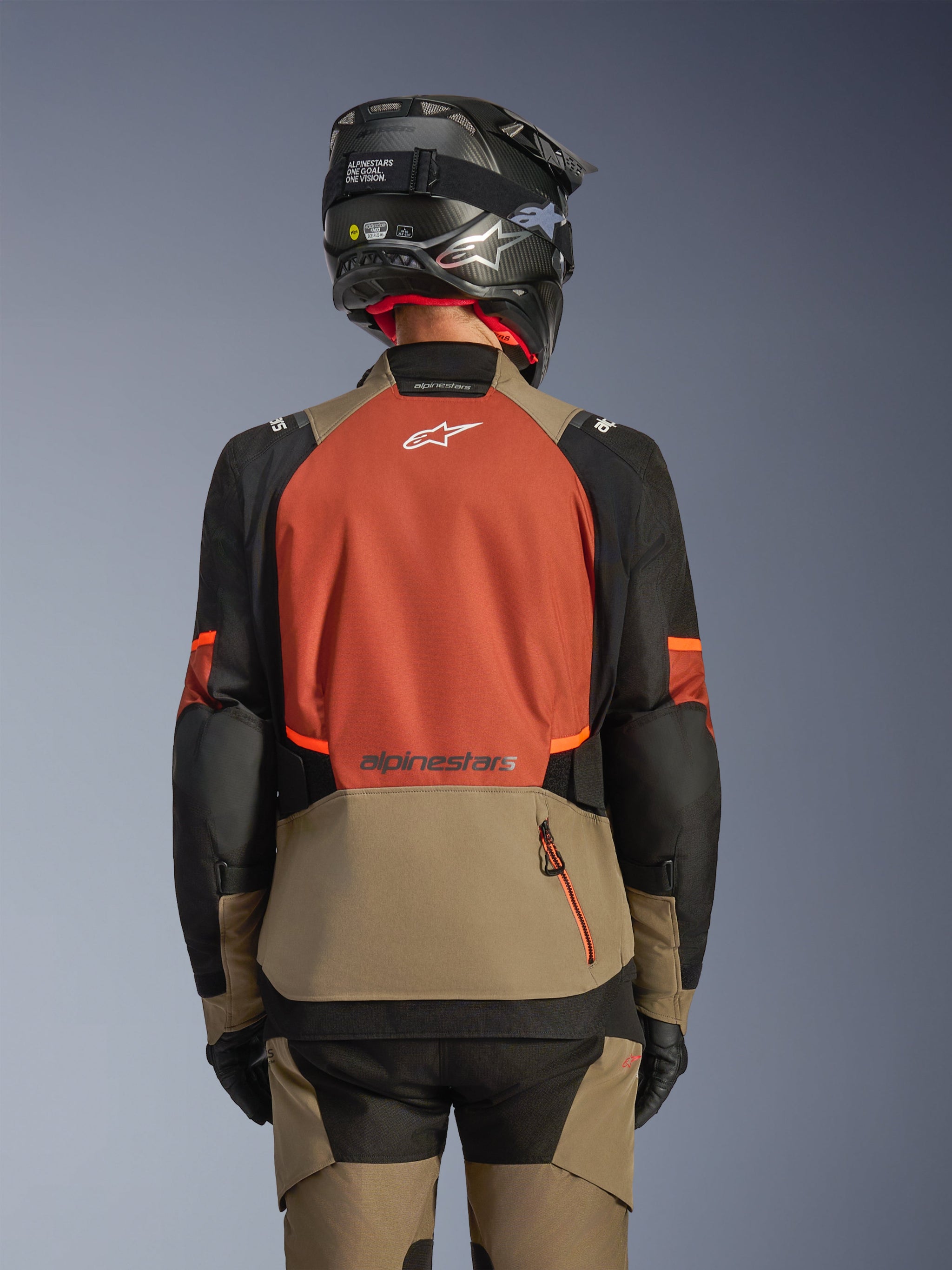 Back view of the Alpinestars Andes V4 Drystar® Jacket in Walnut Rust Black, adventure touring jacket featuring a large rust-colored back panel with a white logo, walnut lower section with a vertical zipper pocket, black sleeves with orange accents, styled with matching pants and a black helmet for all-weather riding.