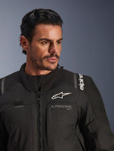 Alpinestars Andes V4 Drystar® Jacket, Black, worn by a male model, close-up shot showing the chest and shoulders, featuring white Alpinestars logos, textile material with ventilation details and hydration hose loop, adventure-touring motorcycle gear