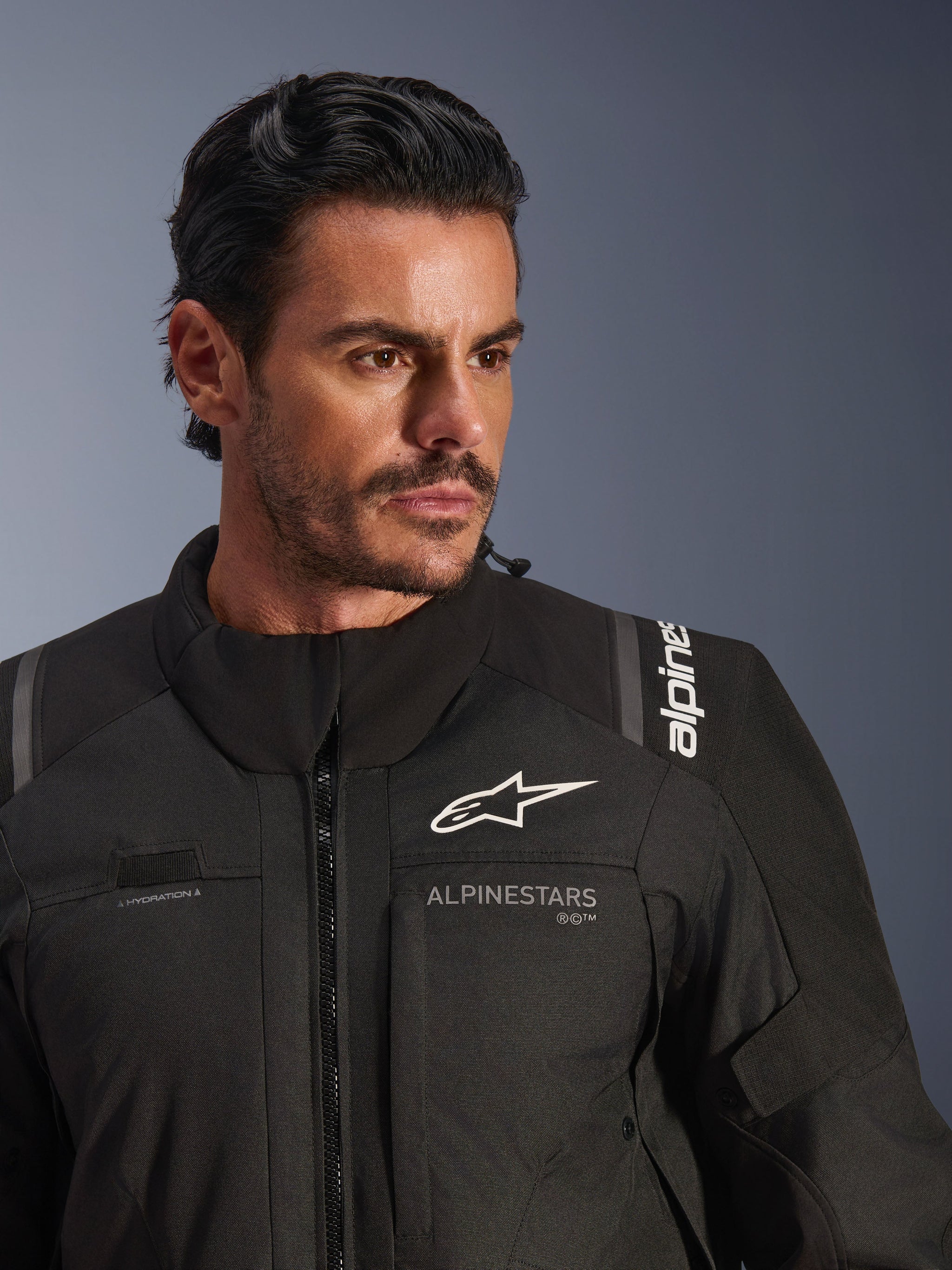 Alpinestars Andes V4 Drystar® Jacket, Black, worn by a male model, close-up shot showing the chest and shoulders, featuring white Alpinestars logos, textile material with ventilation details and hydration hose loop, adventure-touring motorcycle gear