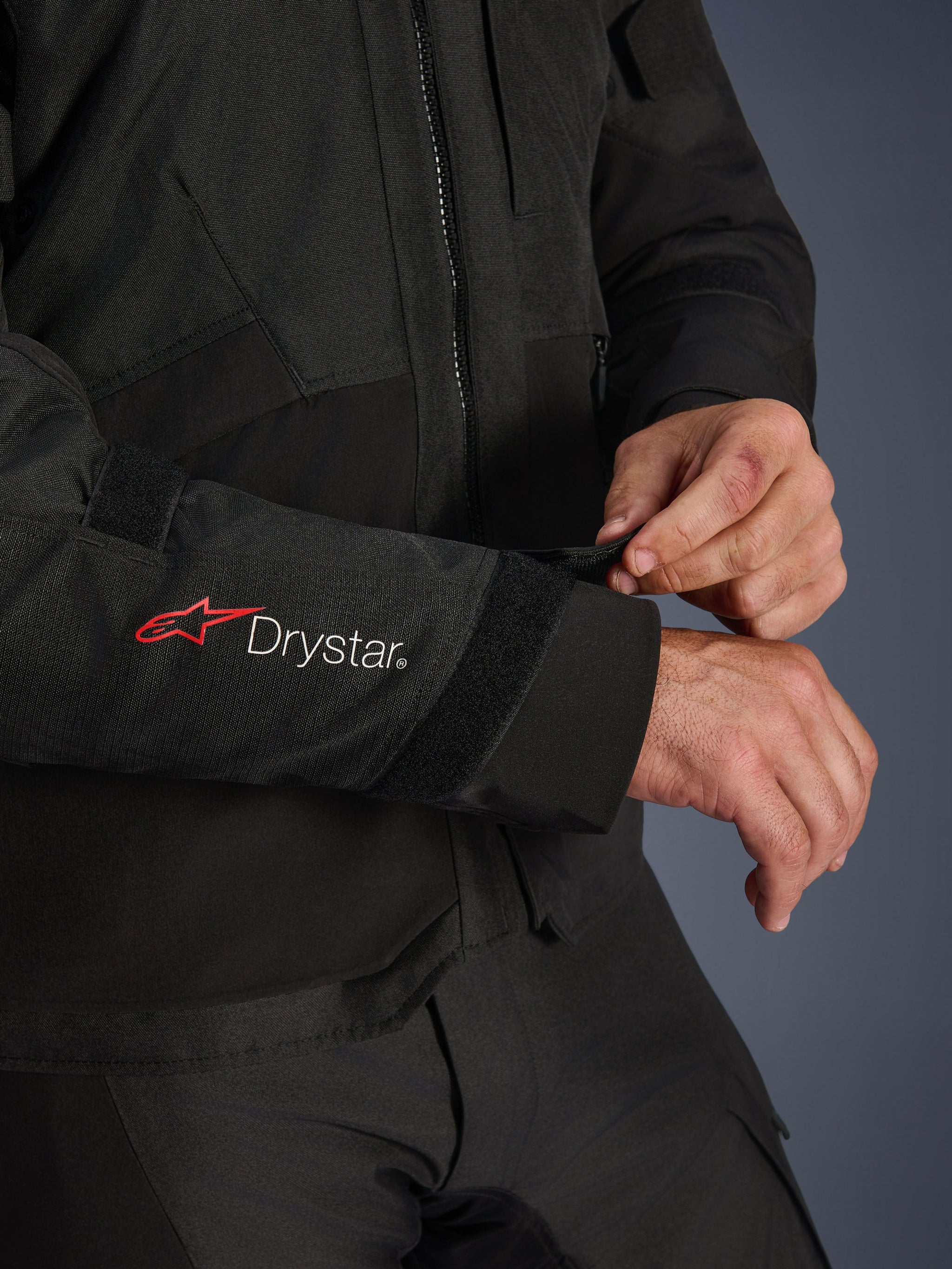 Alpinestars Andes V4 Drystar® Jacket, Black textile motorcycle jacket, close-up of the sleeve featuring red Alpinestars and white Drystar® branding, rider adjusting the velcro wrist strap for a secure fit, adventure riding functional detail