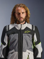 Alpinestars Andes V4 Drystar® Jacket, Dark Gray Ice Gray Black and Yellow Fluo, worn by a man with long wavy hair, featuring technical multi-fabric construction, Alpinestars branding on chest and shoulders, high collar design for adventure riding