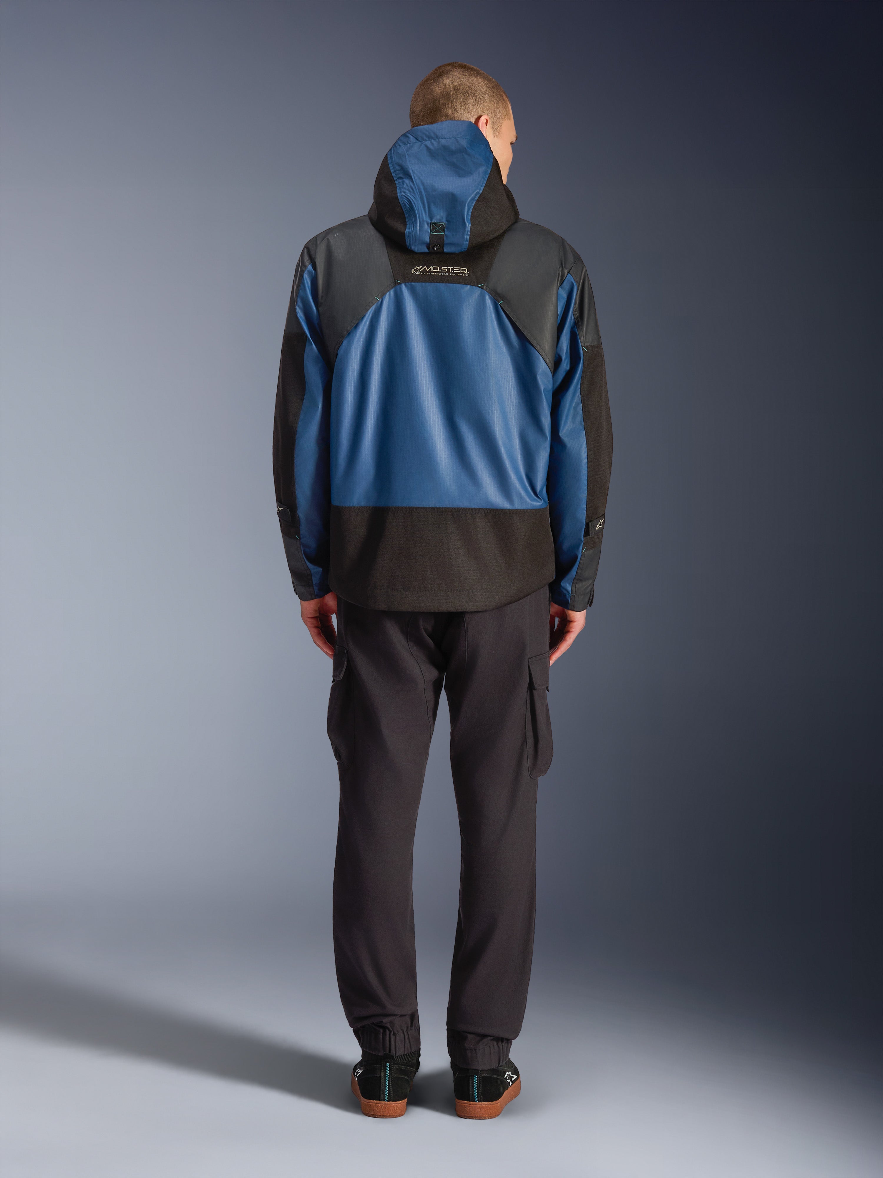 Alpinestars Mospher Waterproof Jacket in Black and Dark Petrol, rear view on model, paired with black cargo pants, featuring tactical ripstop construction and MO.ST.EQ. branding on the back.