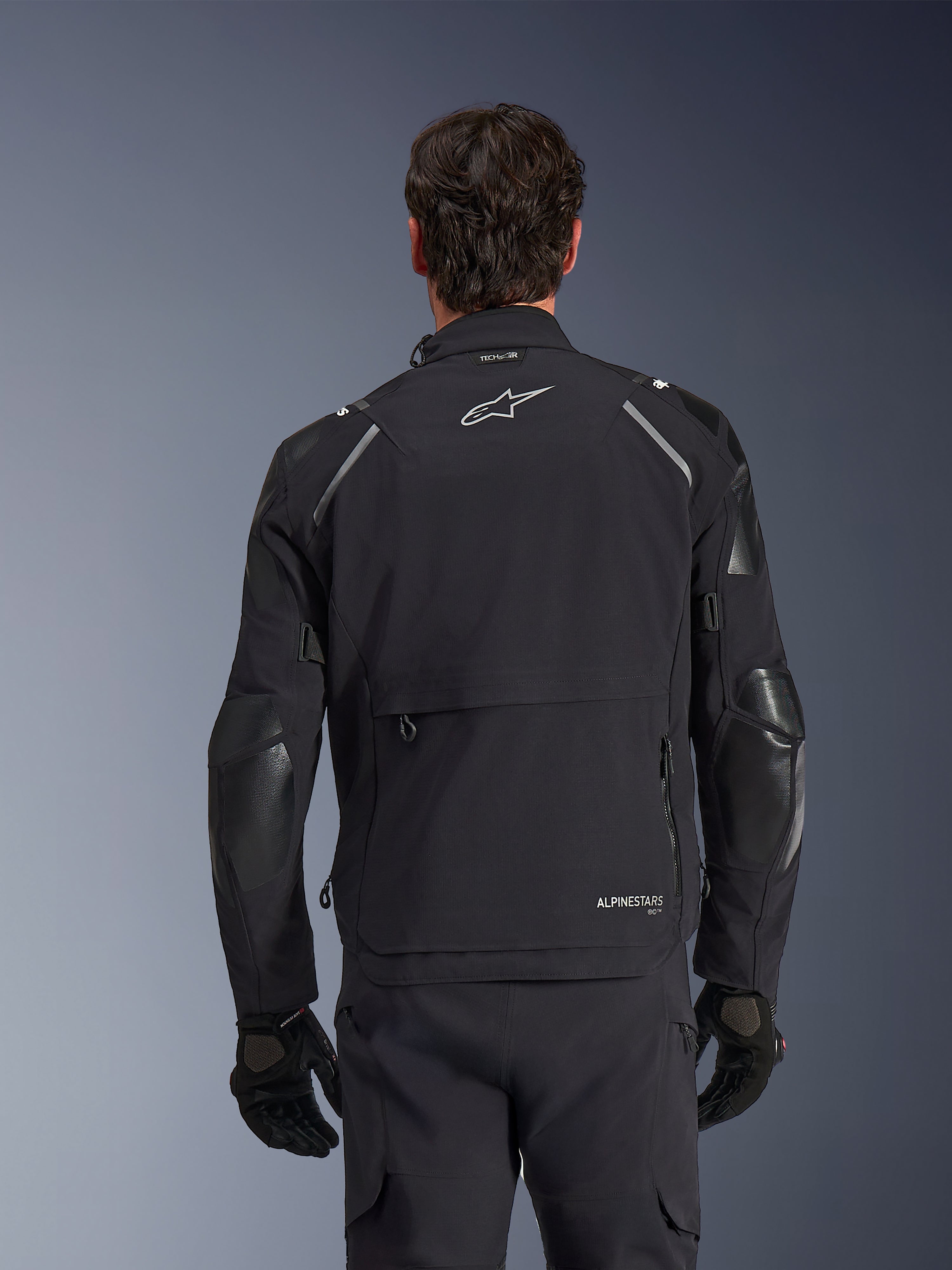 Alpinestars Andes Pro Drystar® XF Jacket, Black and Dark Gray, back view worn by a male model, featuring a large rear storage compartment, reflective shoulder accents, and logo branding, paired with black gloves and pants for adventure touring.