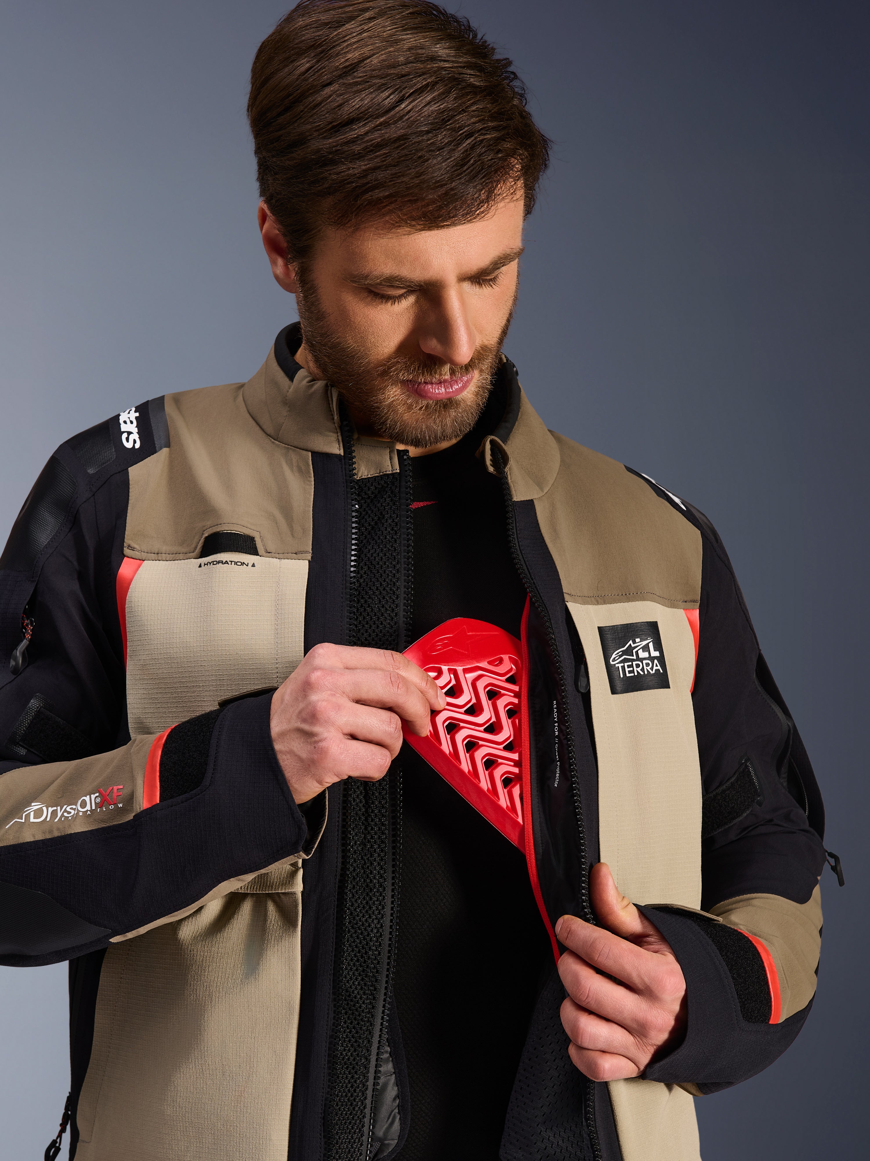 Alpinestars Andes Pro Drystar® XF Jacket in Walnut Aluminum, model demonstrating the red Nucleon chest protector insert into the internal pocket, adventure motorcycle jacket with ALL TERRA branding and Drystar XF logos.