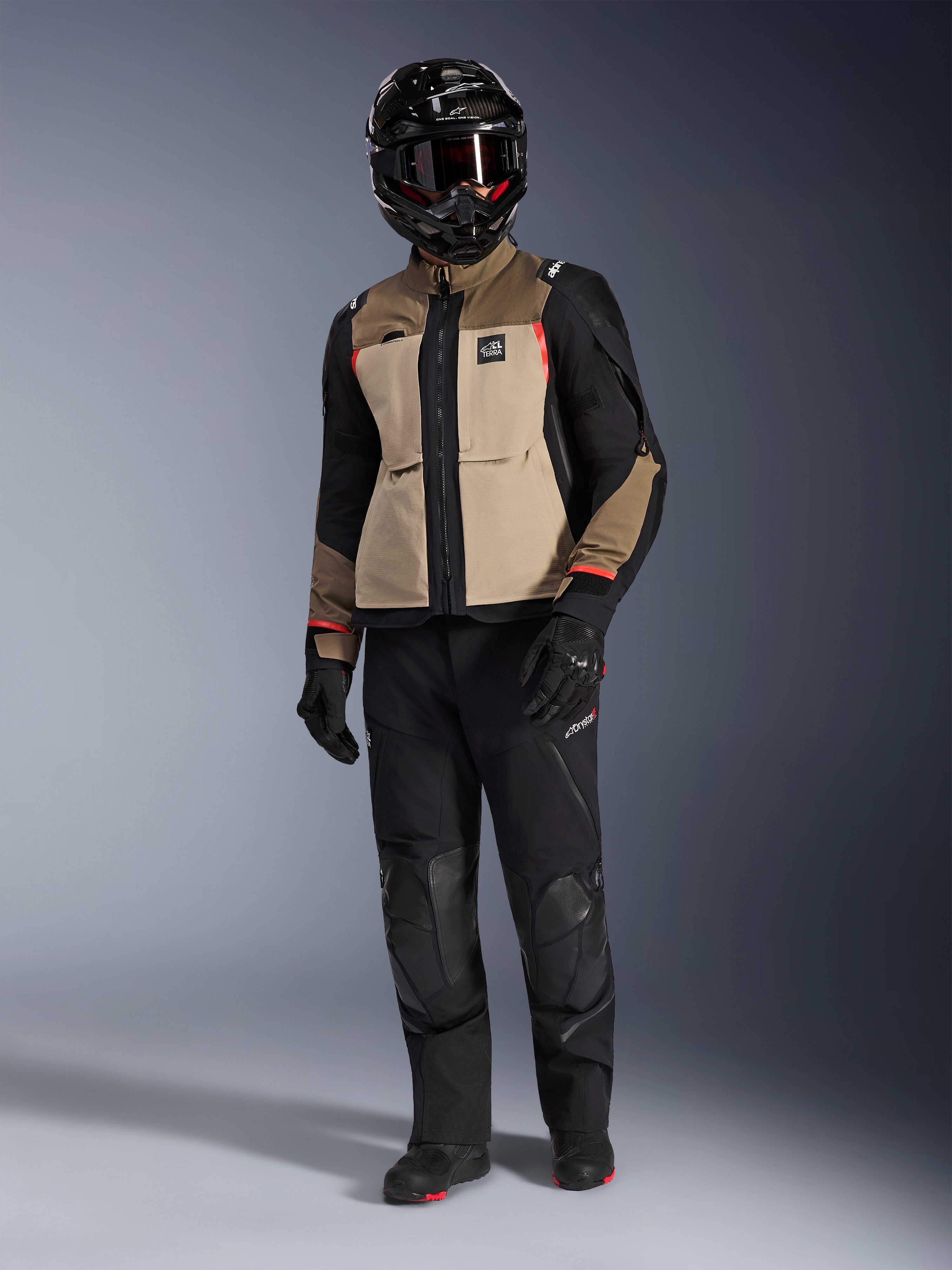 Alpinestars Andes Pro Drystar® XF Jacket in Walnut Aluminum, adventure motorcycle jacket worn with matching black pants, a black adventure helmet with goggles, and black boots, featuring tan and black panels with red accents.