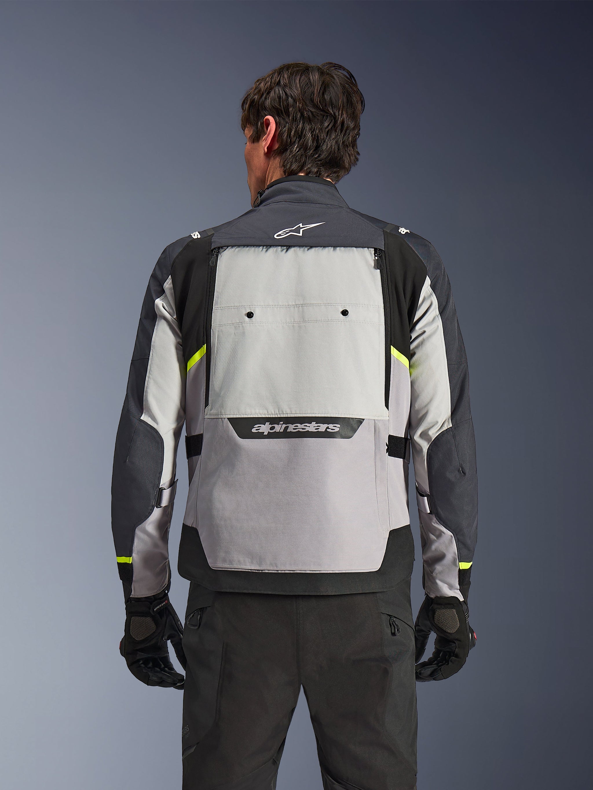 Rear view of a rider wearing the Alpinestars Cusco Drystar® Jacket in Ice Gray, Mid Gray, and Deep Gray, featuring neon yellow accents, a large back ventilation panel, and bold Alpinestars branding, paired with black riding pants and gloves.