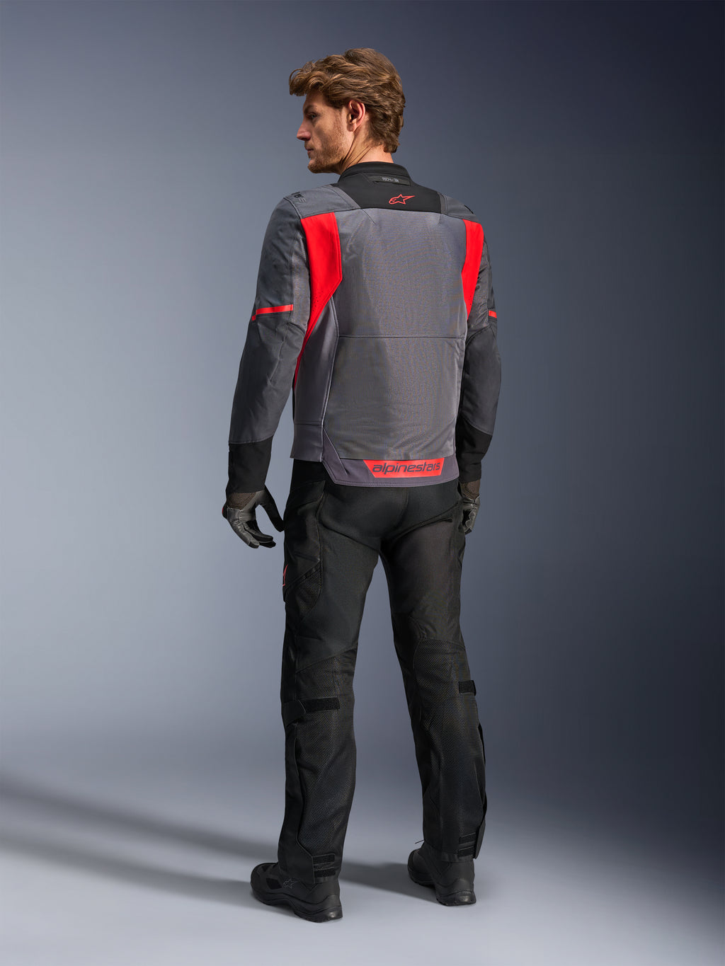 Male rider from behind wearing the Alpinestars St-2 Air Jacket in Deep Gray, Dark Gray and Bright Red, showcasing the large mesh ventilation panel on the back and red branding, styled with black motorcycle pants and gray gloves.
