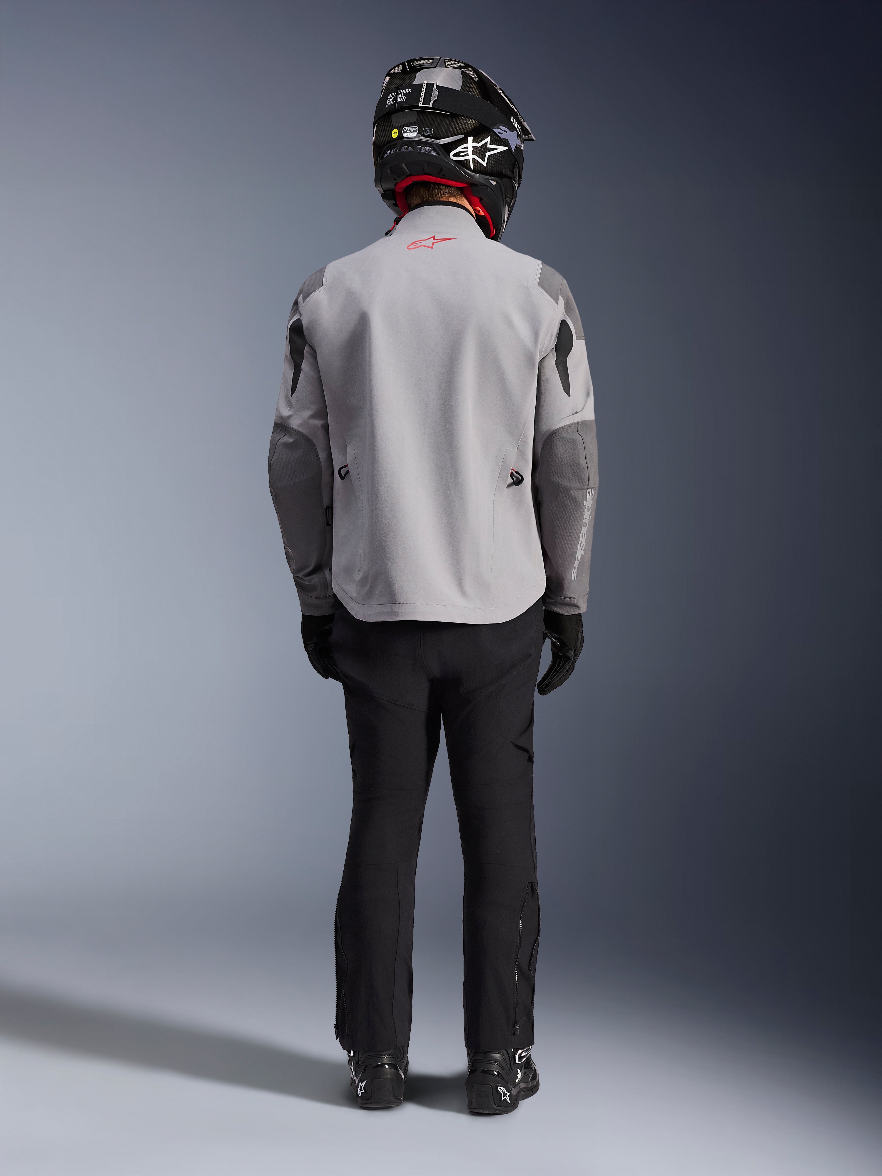 Alpinestars Maxdura Drystar®XF Laminated Armored Shell, motorcycle jacket in Mid Gray and Dark Gray, seen from the back on a rider, worn with black pants, black boots, and a black and white helmet, adventure touring gear