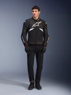 Male model wearing the Alpinestars T-GP R V3 Drystar® Jacket in Black and White, waterproof textile sport jacket with large white Alpinestars logo on the chest, worn with black riding pants and gloves, professional studio portrait highlighting the sleek sport silhouette.