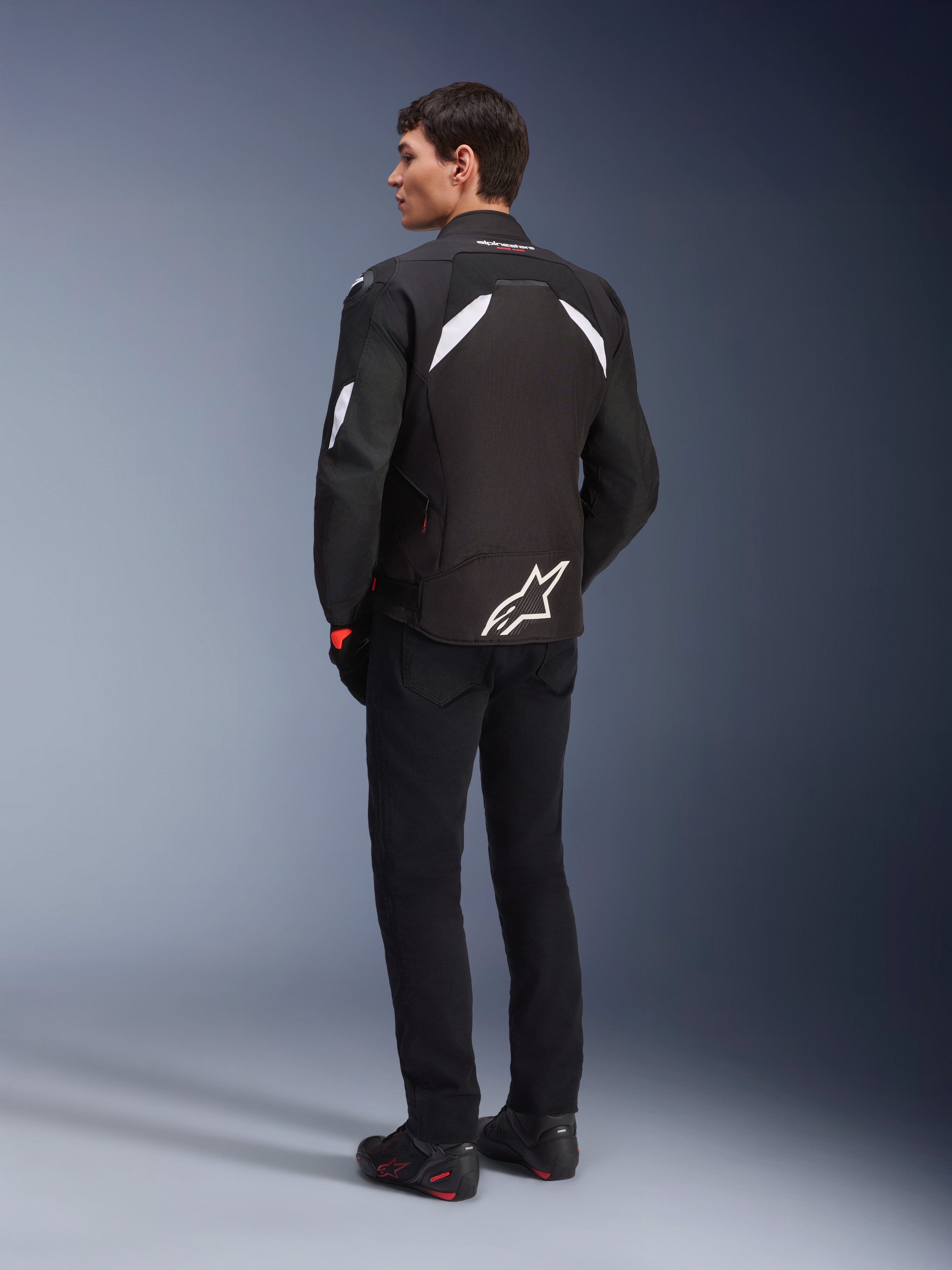 Male model from the back wearing the Alpinestars T-GP R V3 Drystar® Jacket in Black and White, featuring white shoulder accents and a large white logo on the lower back, paired with black riding pants and boots, sport textile motorcycle jacket.