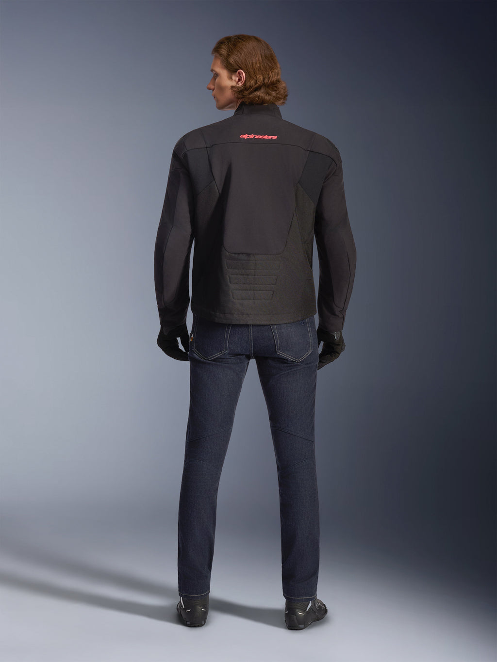 Alpinestars SMX Waterproof Jacket, Black, back view worn by a model, featuring a red logo on the upper back, elongated rear hem for riding coverage, styled with dark blue motorcycle jeans and black gloves.