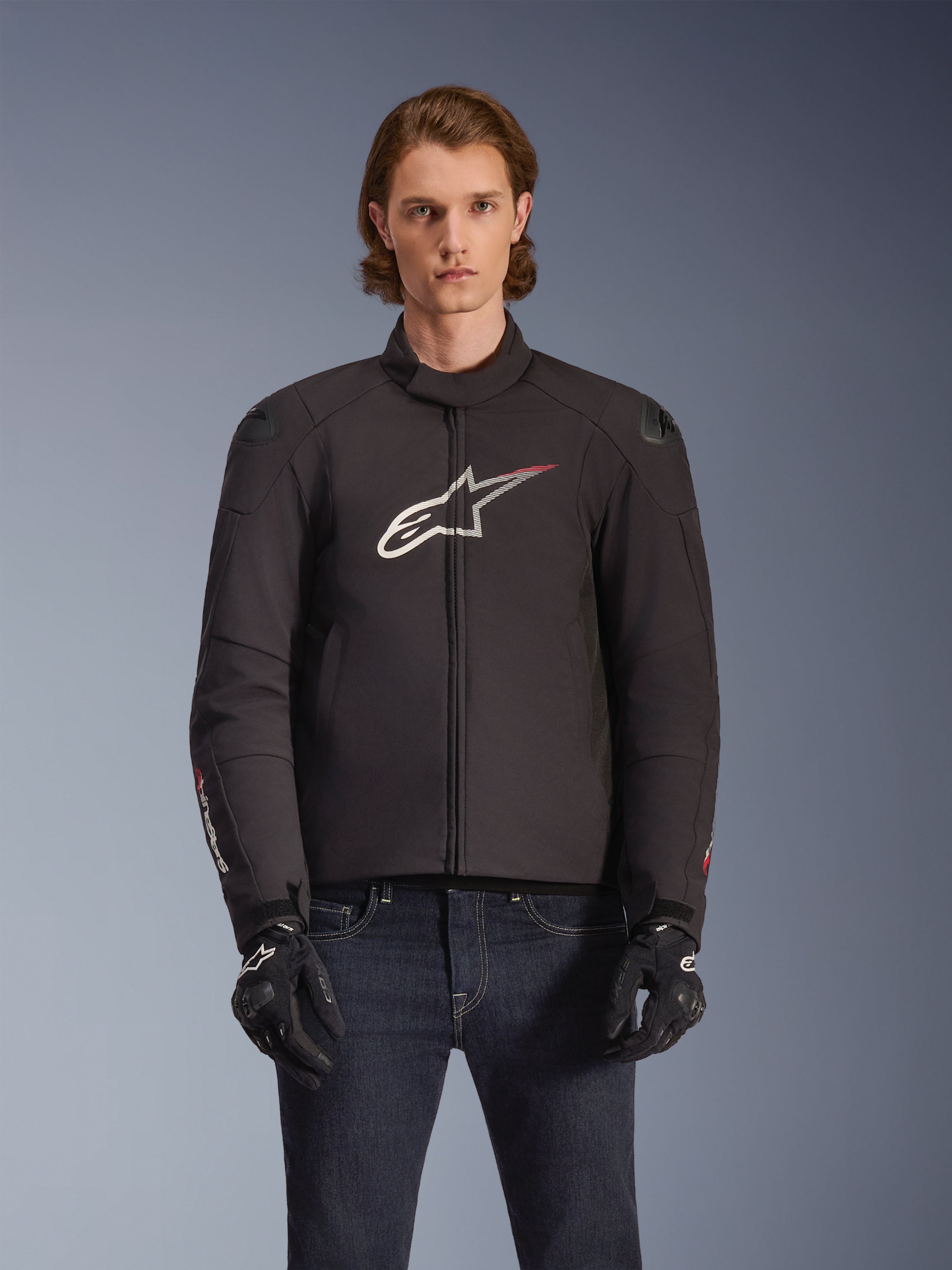 Alpinestars SMX Waterproof Jacket, Black textile motorcycle jacket with white and red star logo on the chest, worn by a model with black gloves and blue jeans, featuring external shoulder sliders and a sporty fit.