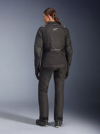 Female rider from behind wearing the Alpinestars Stella Andes V4 Drystar® Jacket, Black, paired with matching motorcycle pants, white Astar logo on the back, elongated rear hem, complete adventure-touring riding outfit including black gloves and boots