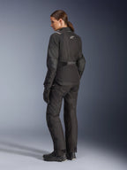 Woman rider wearing Alpinestars Stella Andes V4 Drystar® Jacket, Black adventure motorcycle jacket, 3/4 rear view, paired with matching textile pants and gloves, featuring white Alpinestars logos on the back and waist, reinforced protective panels, and an elongated rear hem.