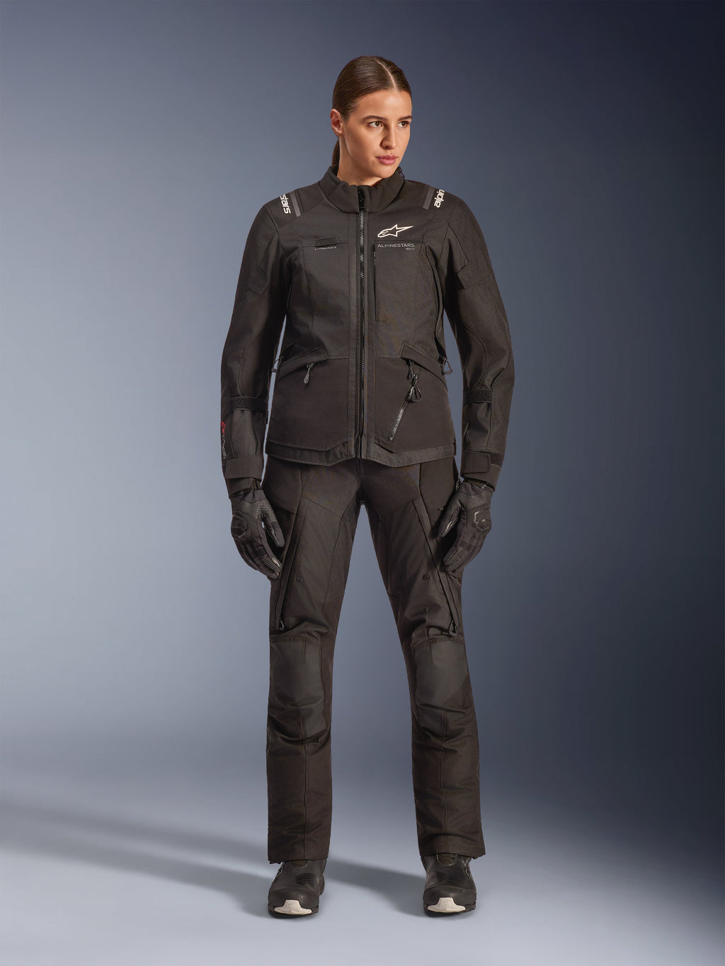 Female rider wearing Alpinestars Stella Andes V4 Drystar® Jacket in Black, paired with matching black adventure-touring pants and gloves, featuring technical textile construction with multiple utility pockets and white Alpinestars branding, complete all-weather motorcycle gear