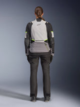 Woman rider from the back wearing the Alpinestars Stella Andes V4 Drystar® Jacket, Ice Gray, Dark Gray, and Black with Yellow Fluo accents, featuring a large rear storage pocket and Alpinestars logo, paired with matching black adventure pants, all-weather motorcycle touring gear