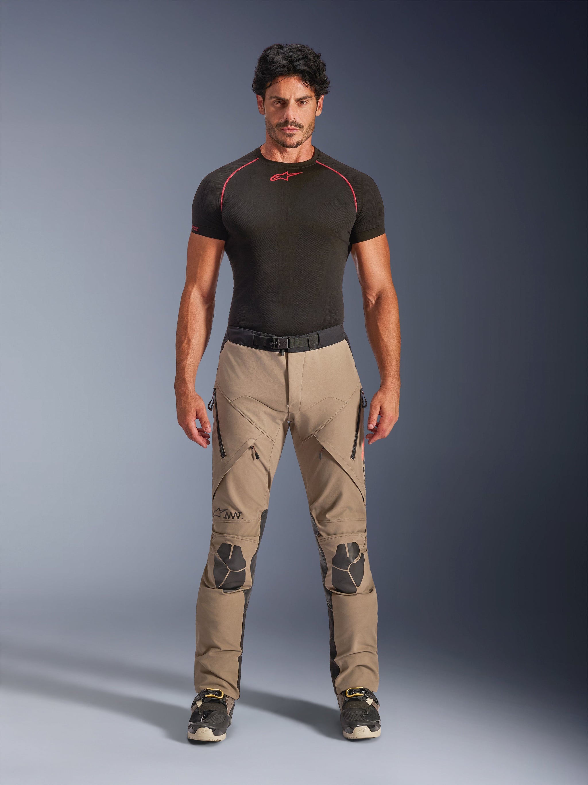 Male model wearing Alpinestars AMT-8 Stretch Drystar® XF Pants in Walnut and Black, paired with a black short-sleeve base layer, adventure touring pants featuring reinforced knees, zippered ventilation, and ergonomic stretch design.
