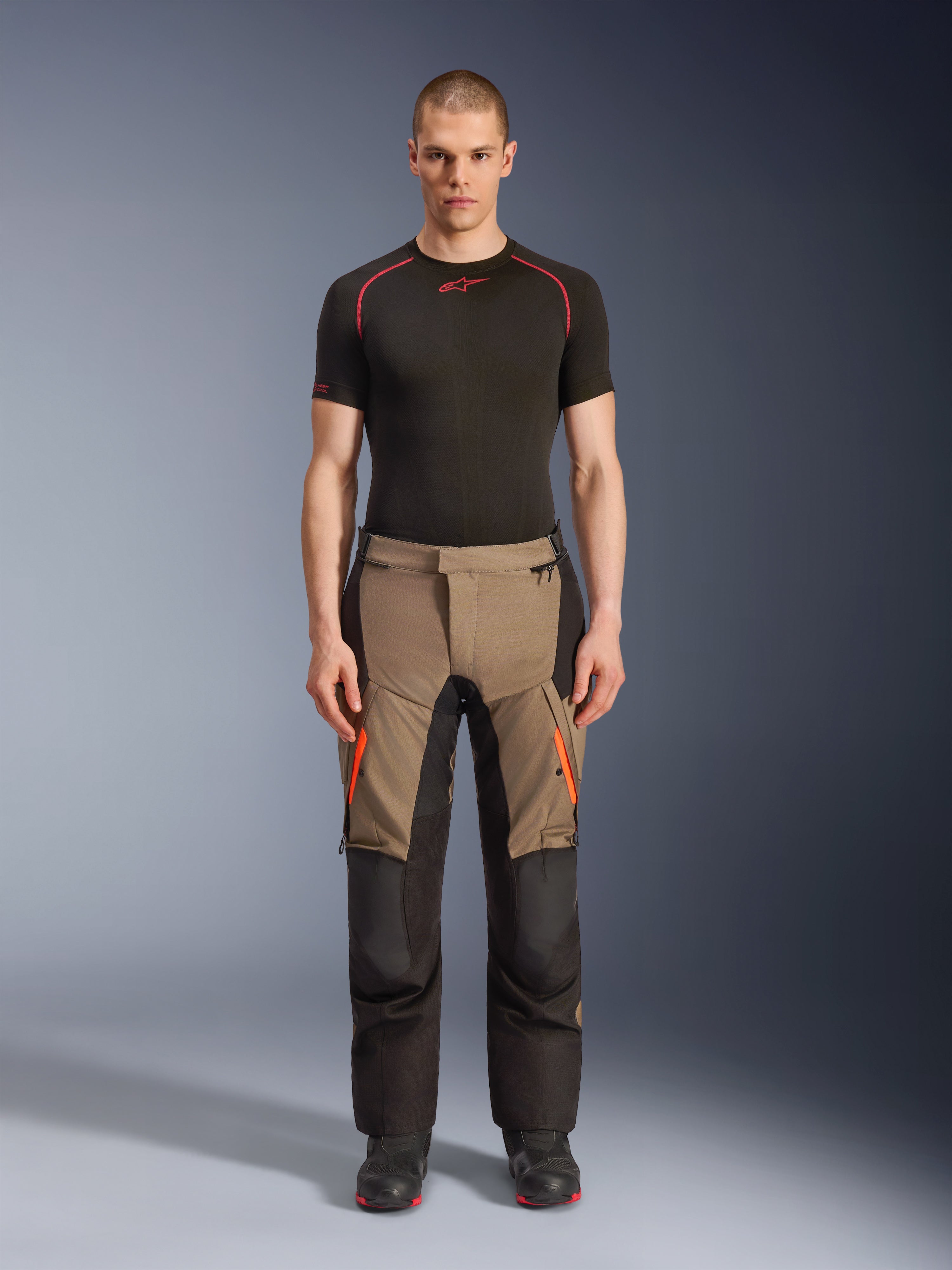 Male model wearing Alpinestars Andes V4 Drystar® Pants in Walnut Black, adventure motorcycle pants paired with a black base layer and boots, featuring orange zipper accents on the thighs and reinforced black panels for all-terrain durability.