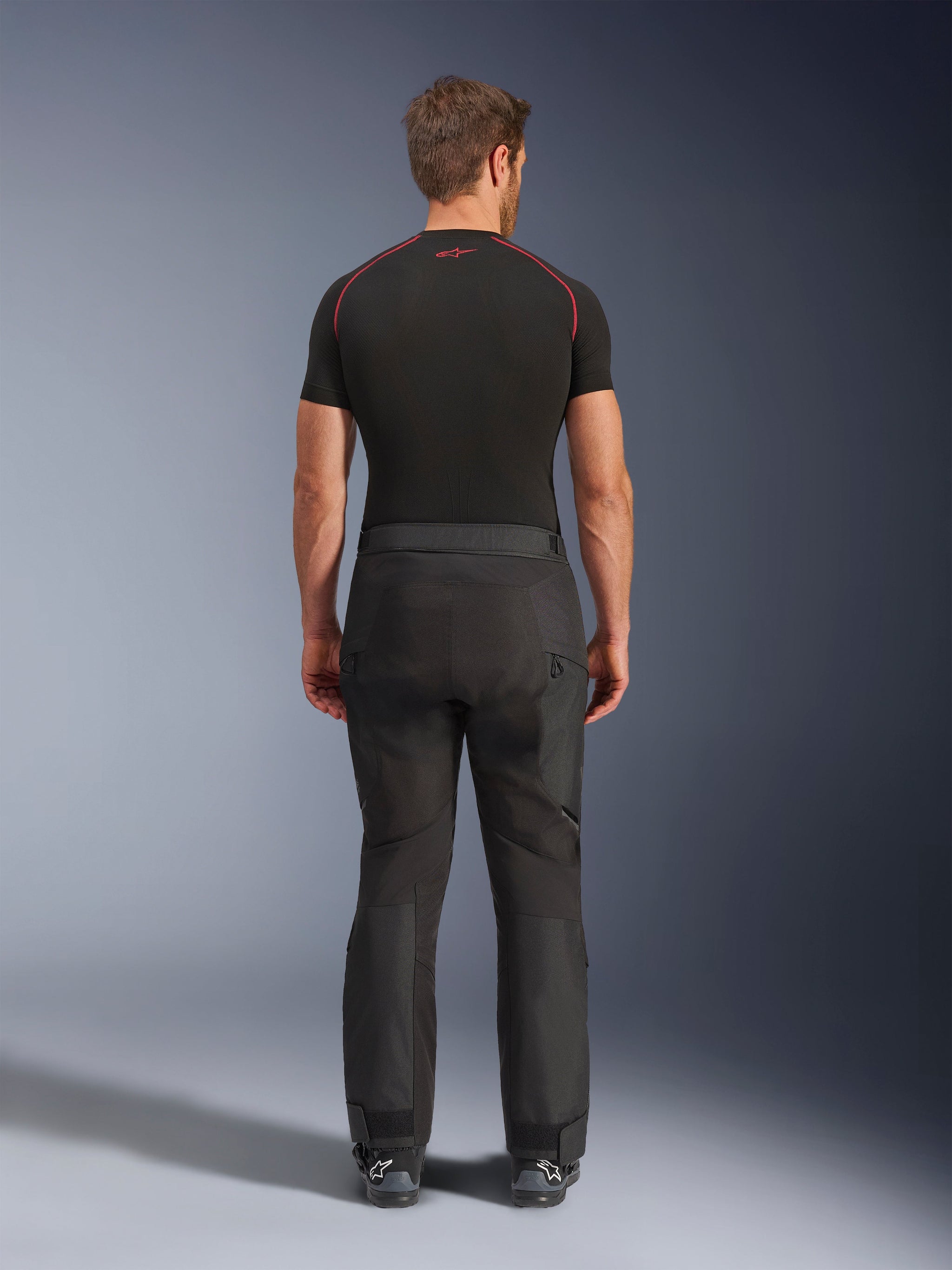 Rider wearing Alpinestars Cusco Drystar® Pants, Black adventure motorcycle pants, back view, paired with a black short-sleeve compression shirt and black riding boots, featuring a durable textile design with articulated panels for touring