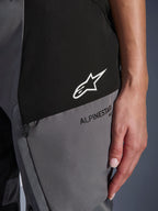 Close-up of Alpinestars Woman Stella Andes V4 Drystar® Pants in Black and Dark Gray, featuring a white star logo on a black panel and black Alpinestars branding on a gray panel, showing technical textile fabric and zippered pocket detail.