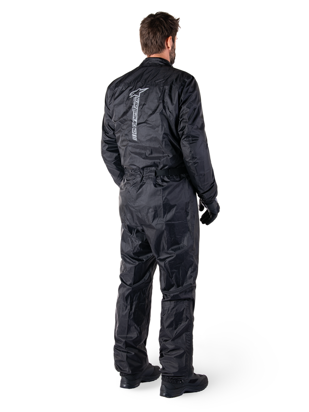 Alpinestars Hurricane Rain V2 Suit, Black one-piece waterproof motorcycle rain suit, back view with large vertical reflective logo, lightweight wind-resistant fabric, worn with black gloves and boots