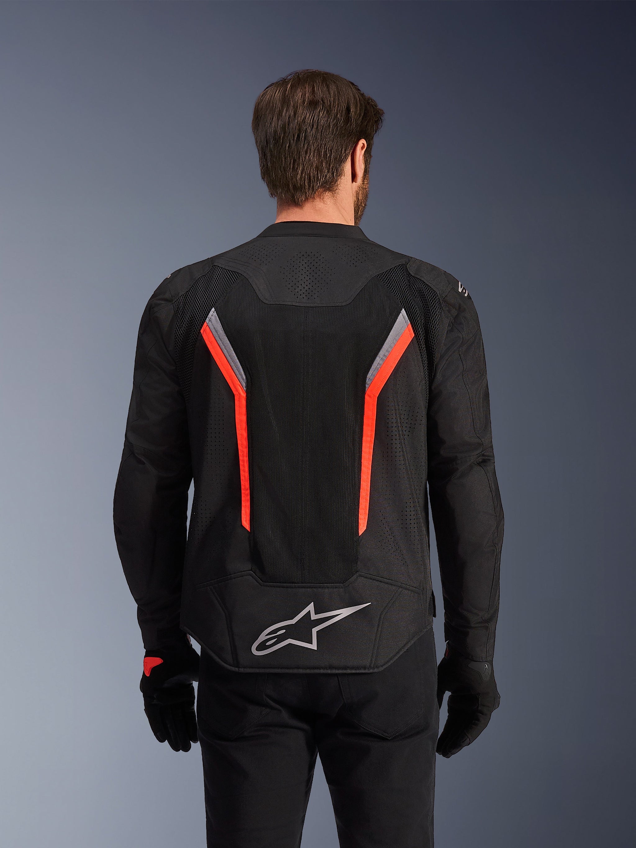 Alpinestars AST V3 Air Jacket, Rear view, Black with Dark Gray and Red Fluo, featuring large mesh back panel and laser perforations, worn with black gloves and pants, sporty design with Alpinestars logo, technical warm-weather motorcycle gear