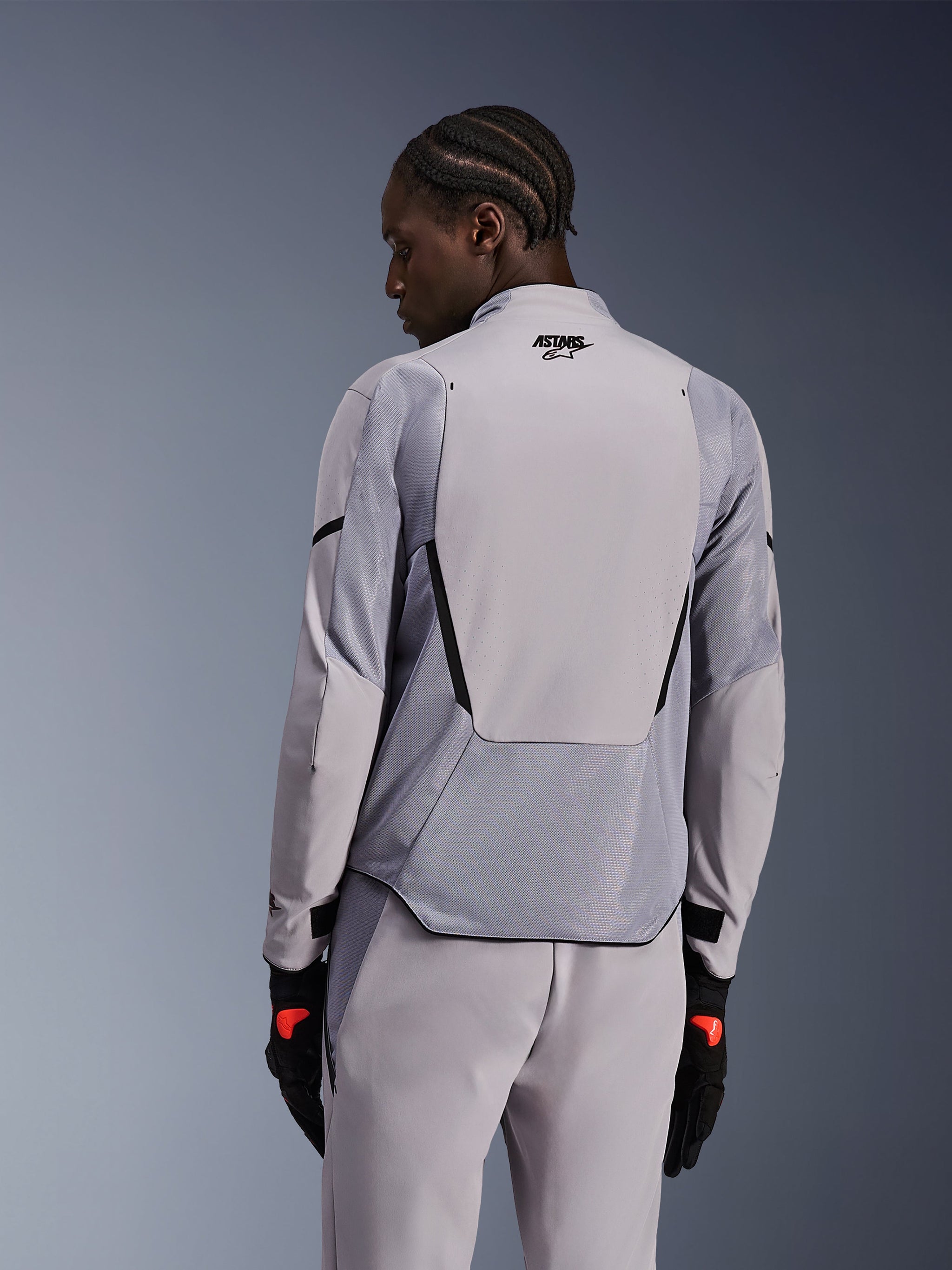 Alpinestars Aeroshell Airflow Jacket in Mid Gray Anthracite, back view showing breathable mesh panels and laser perforations, technical stretch softshell construction with an elongated rear panel, worn with matching gray pants and black gloves for a sport-oriented look.