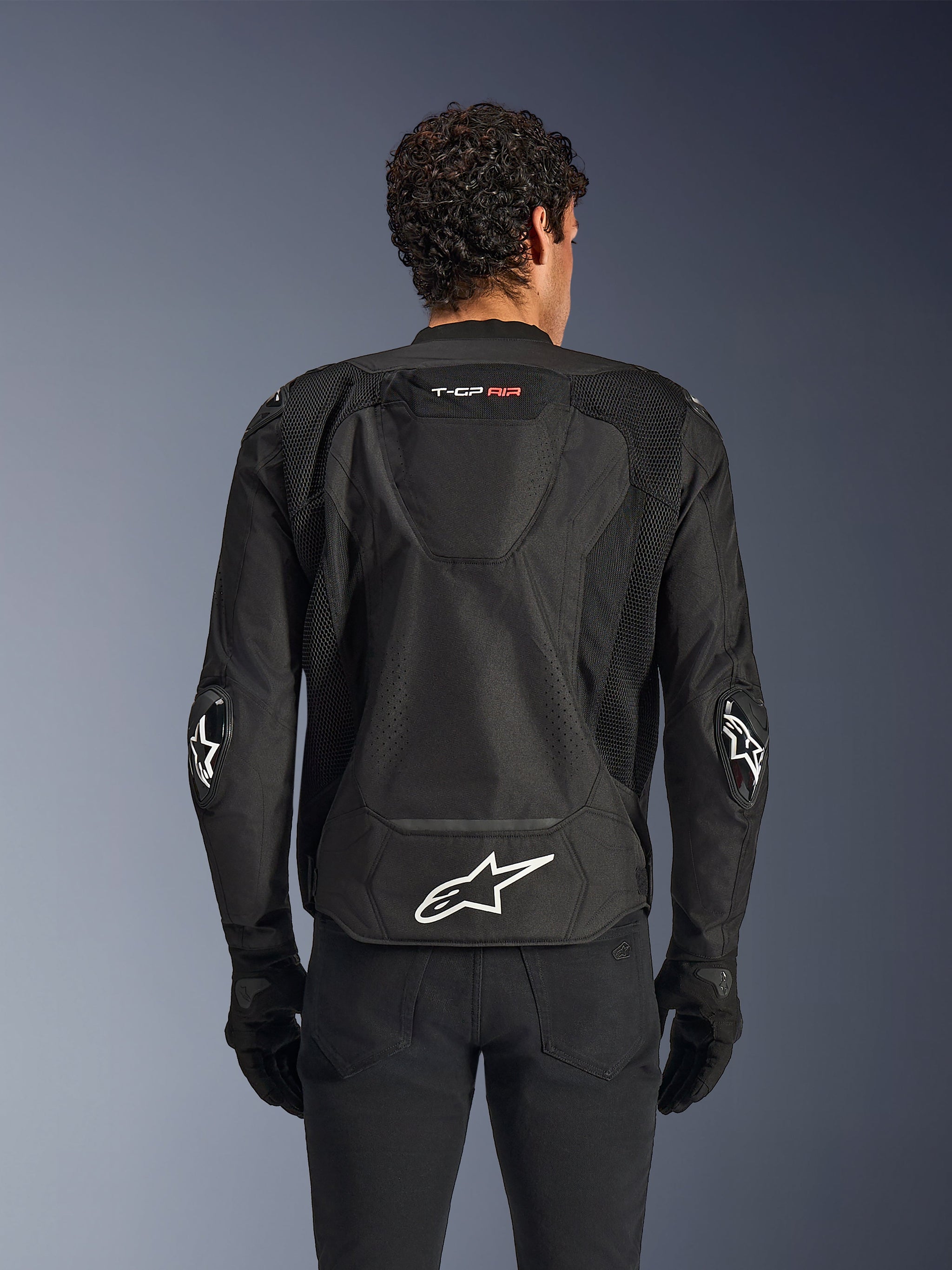 Alpinestars T-GP Air Jacket, Black, rear view of textile motorcycle jacket, featuring an aerodynamic hump, large mesh ventilation panels, and white Alpinestars logo on the lower back, styled with black motorcycle gloves and pants