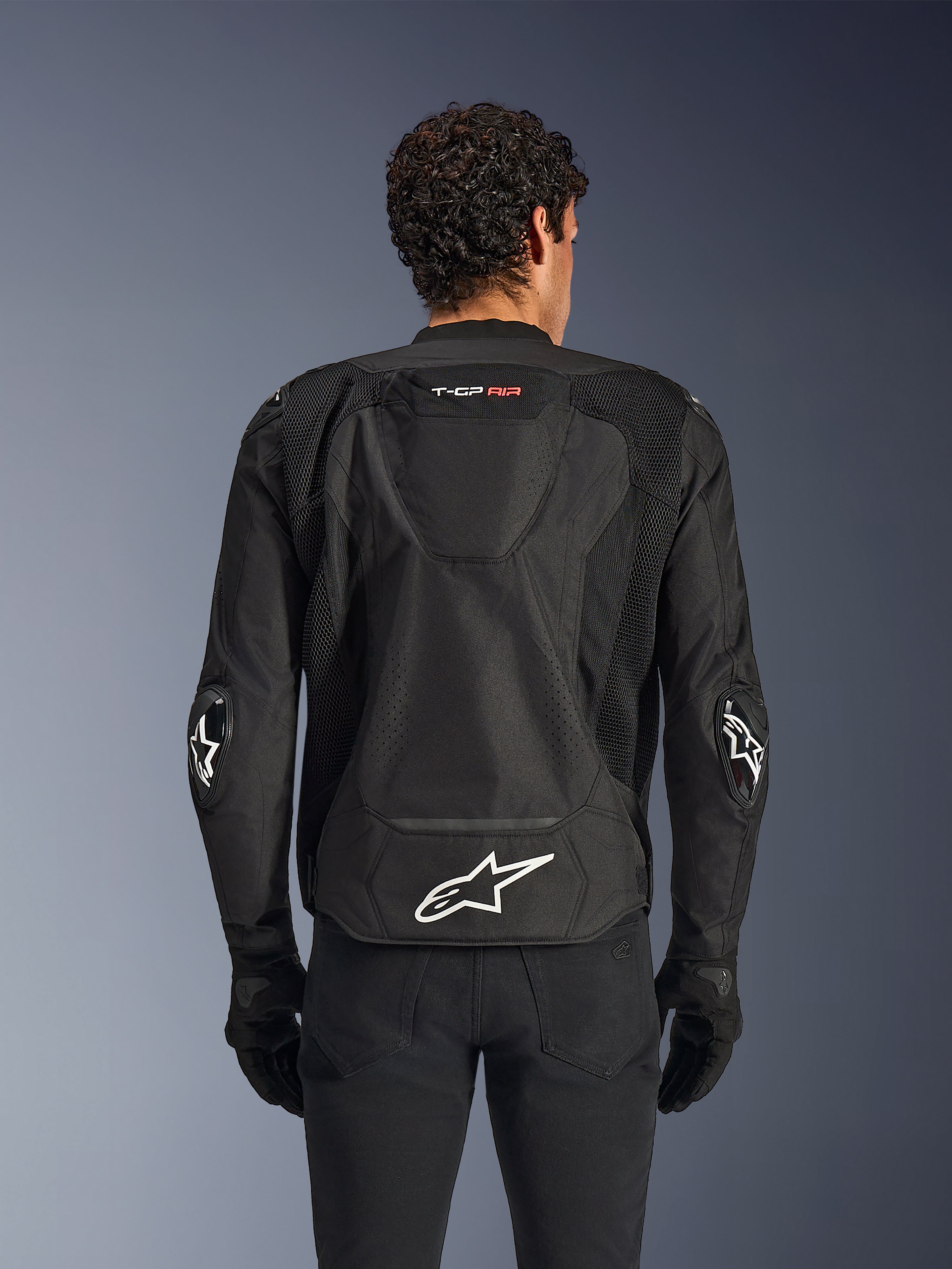 Alpinestars T-GP Air Jacket, Black, rear view of textile motorcycle jacket, featuring an aerodynamic hump, large mesh ventilation panels, and white Alpinestars logo on the lower back, styled with black motorcycle gloves and pants
