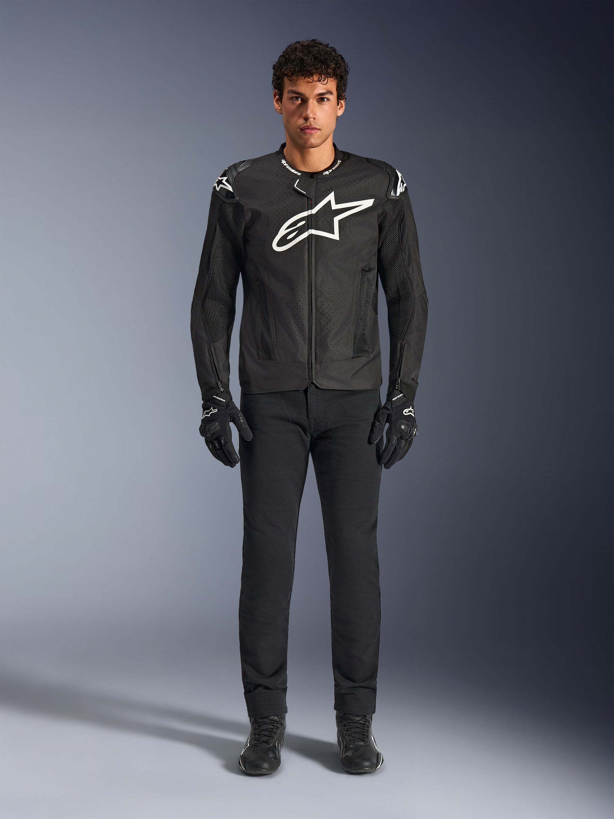 Alpinestars T-GP Air Jacket, Black textile motorcycle jacket with a large white Astars logo on the chest, worn by a model with black riding pants and gloves, featuring perforated panels and mesh ventilation for sport riding