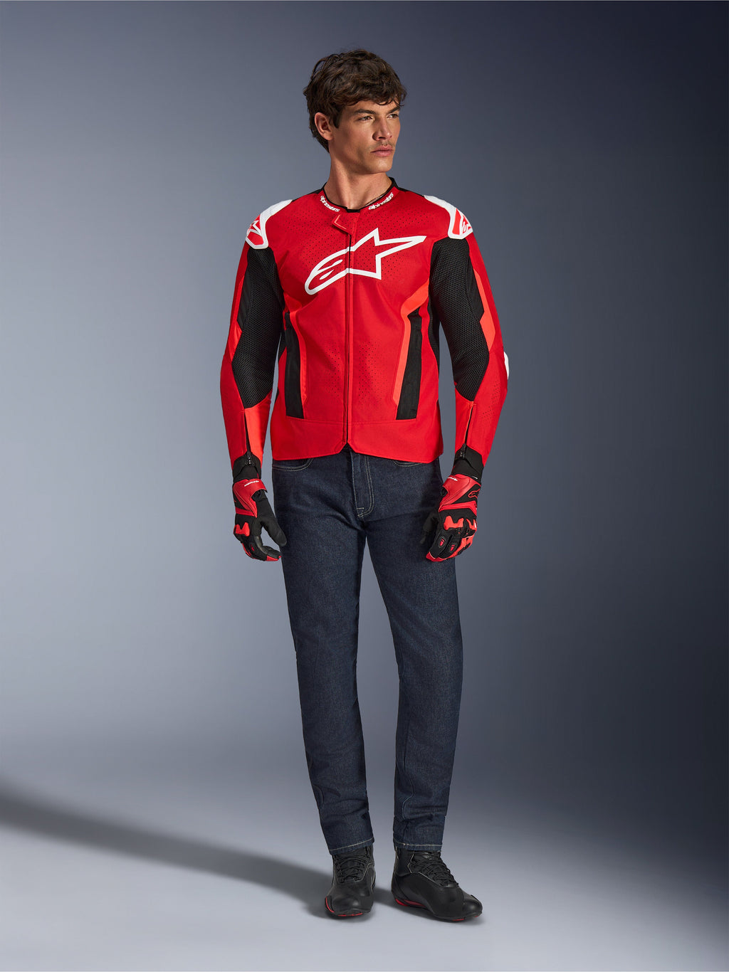 Alpinestars T-GP Air Jacket, Bright Red Red Fluo Black, male model wearing full motorcycle outfit with dark blue jeans and matching gloves, sport textile jacket featuring perforated mesh panels and white Astars logo, studio shot.