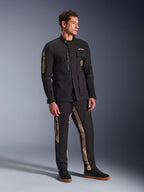Male model wearing Alpinestars Dice Jacket in Black, urban motorcycle jacket with vintage field jacket aesthetic, featuring multiple utility pockets and gold branding on the sleeve, paired with matching black and tan pants and black sneakers.
