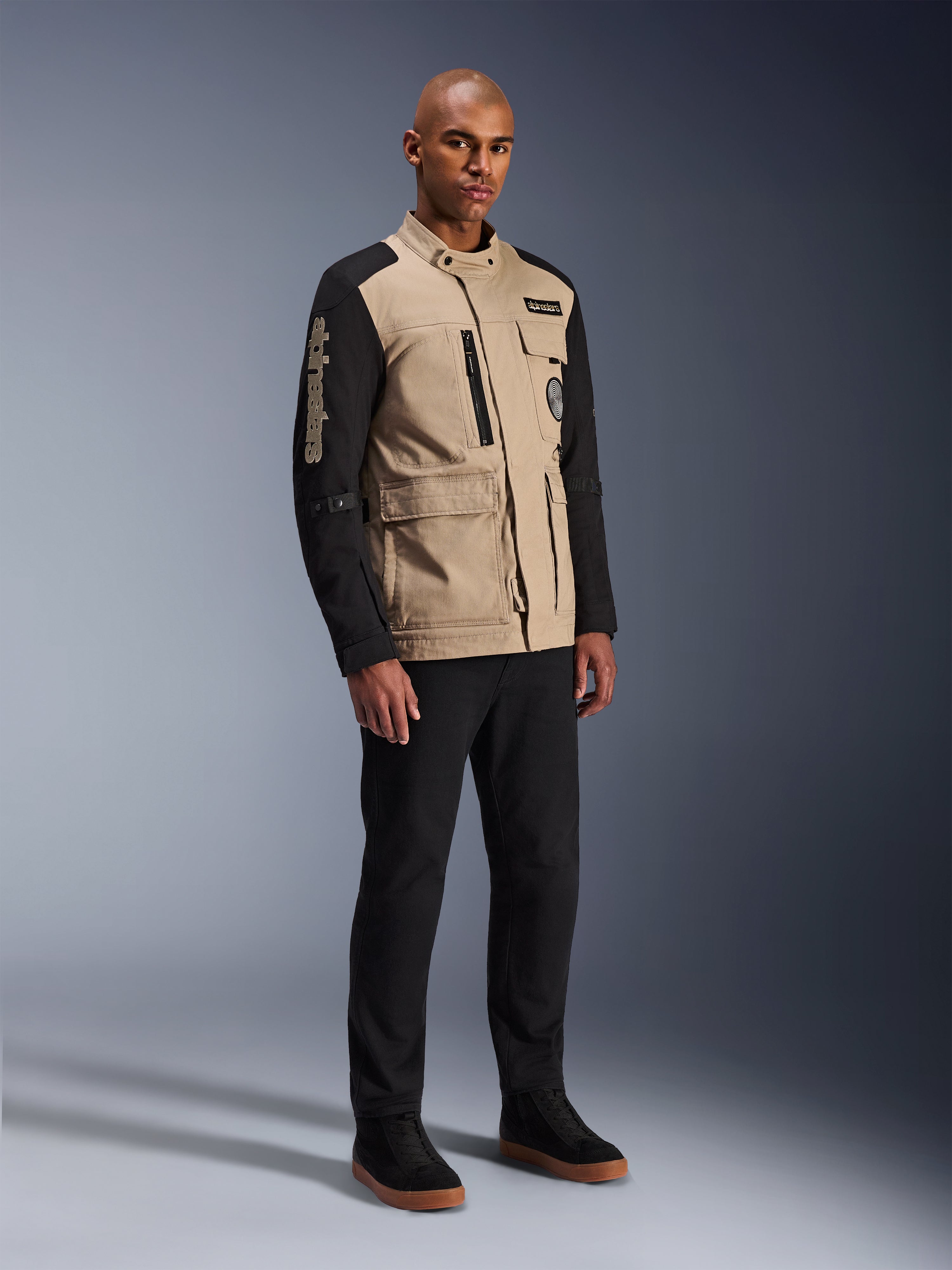 Alpinestars Dice Jacket, Urban Motorcycle Jacket, Aluminum Black and Tan, two-tone design with black sleeves and a khaki torso, worn by a model with black pants and black sneakers, workwear-inspired aesthetic with utility pockets and embroidered branding on the sleeve.